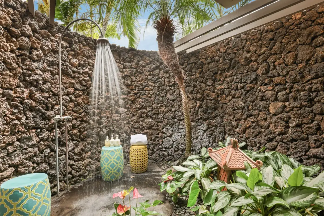 An outdoor shower stands among tropical plants and rough stone walls.