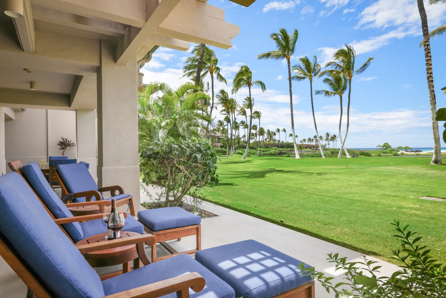 A covered patio with cushioned seating overlooks a palm-lined lawn and ocean.