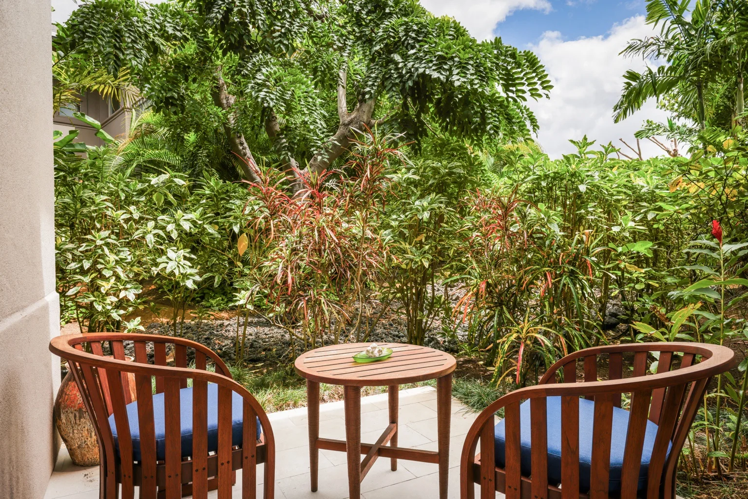 A small outdoor patio with two wooden seats and a round table faces lush greenery.