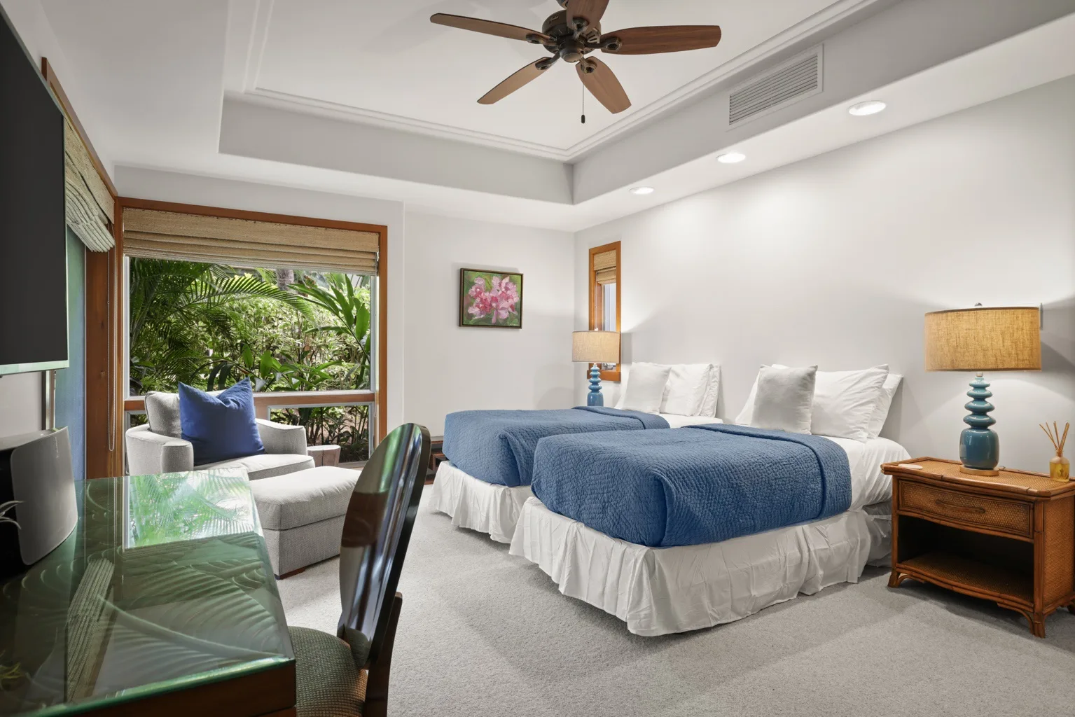 A bright bedroom with a blue-covered bed, seating, and a large window overlooking greenery.