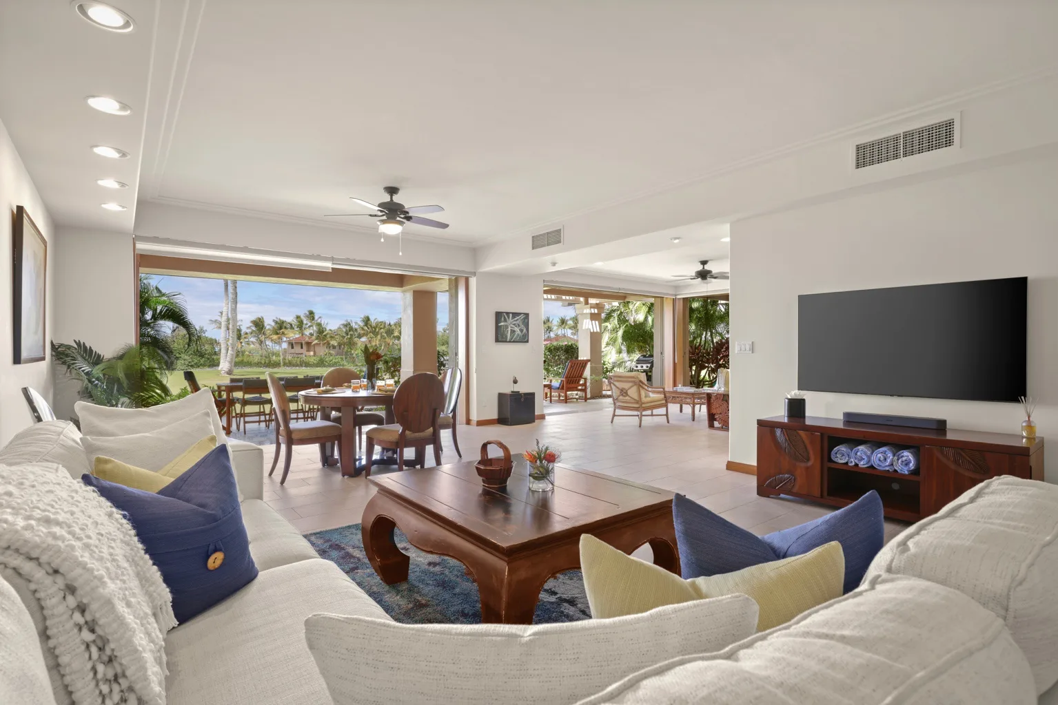 A bright living room opens onto a dining area, with large windows facing greenery.
