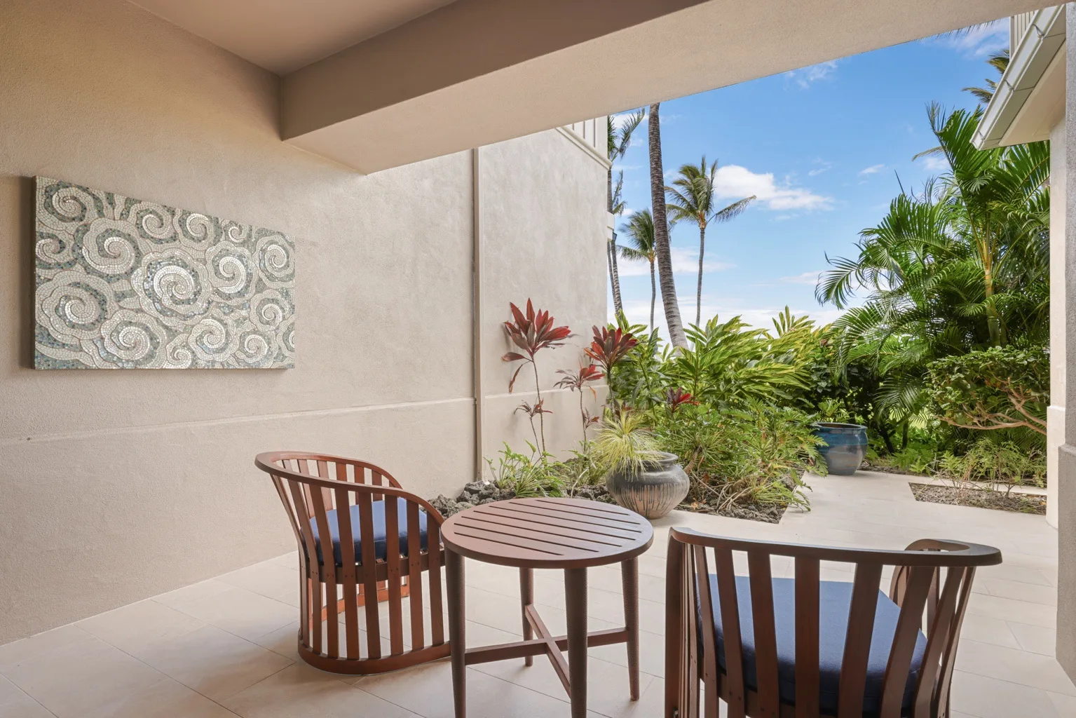 A covered patio has seating and a small table overlooking a tropical garden.