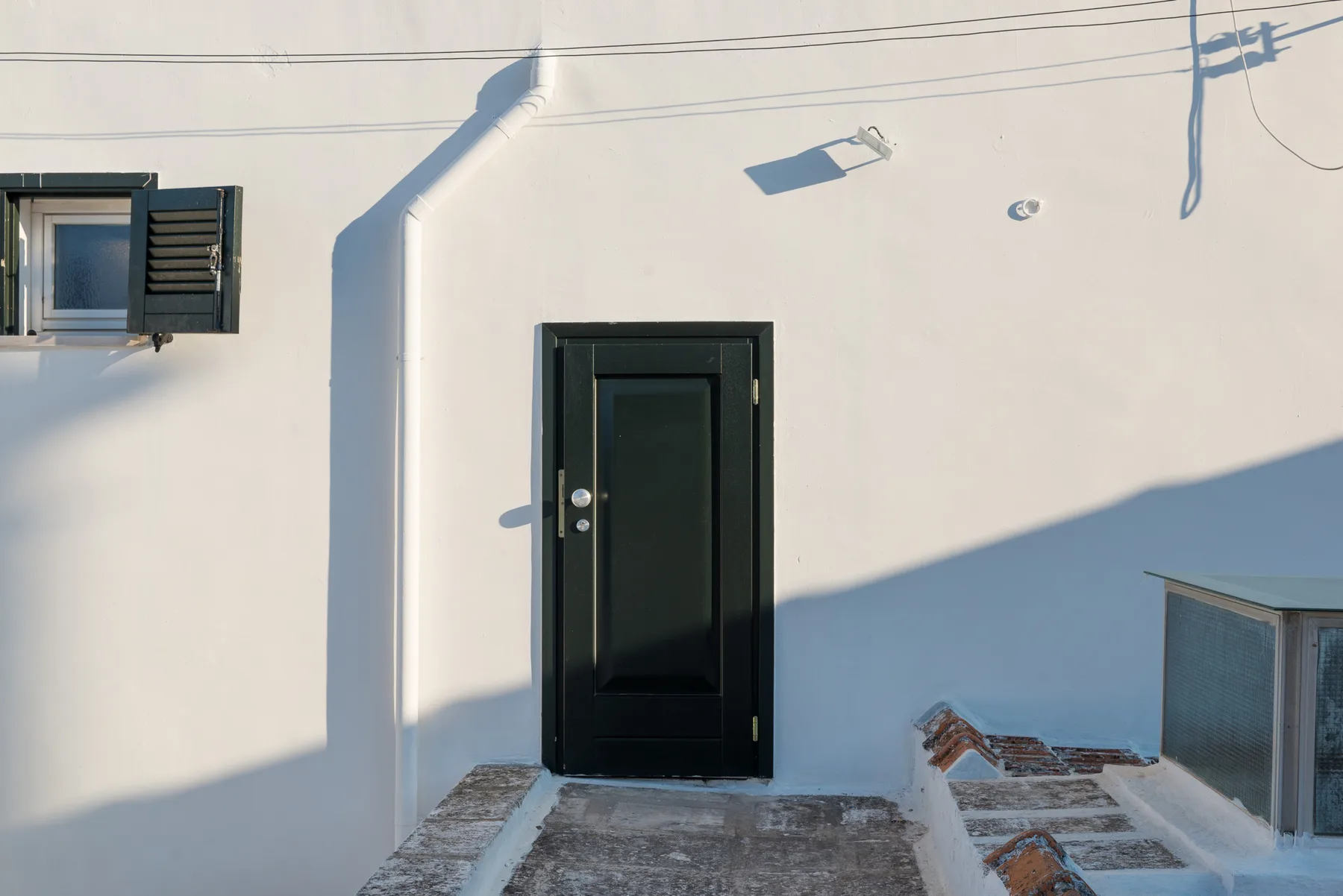 Una puerta negra de entrada está en una pared exterior blanca junto a una ventana pequeña.