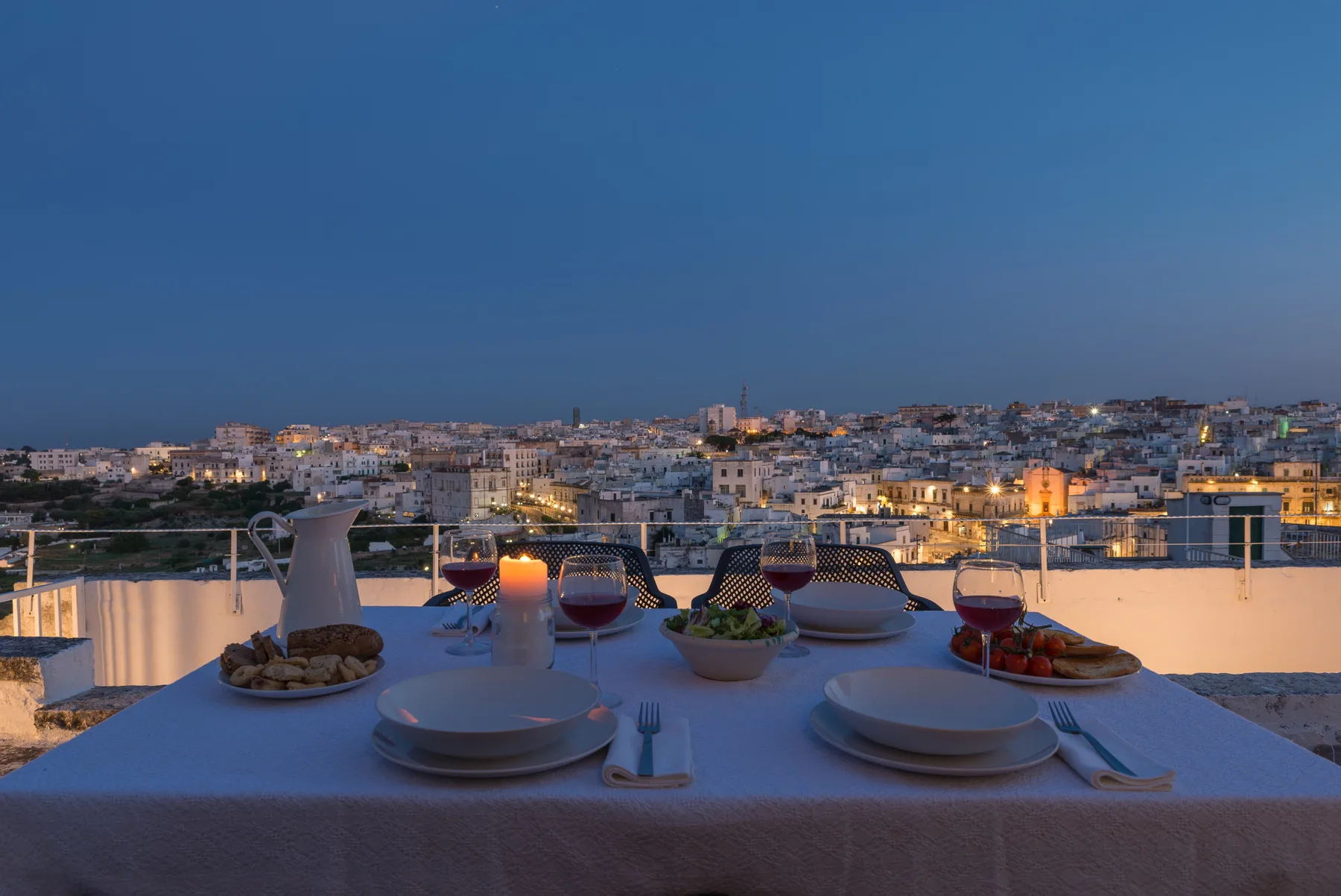 Una mesa de comedor en una terraza ofrece vistas de la ciudad al anochecer, con platos y copas.