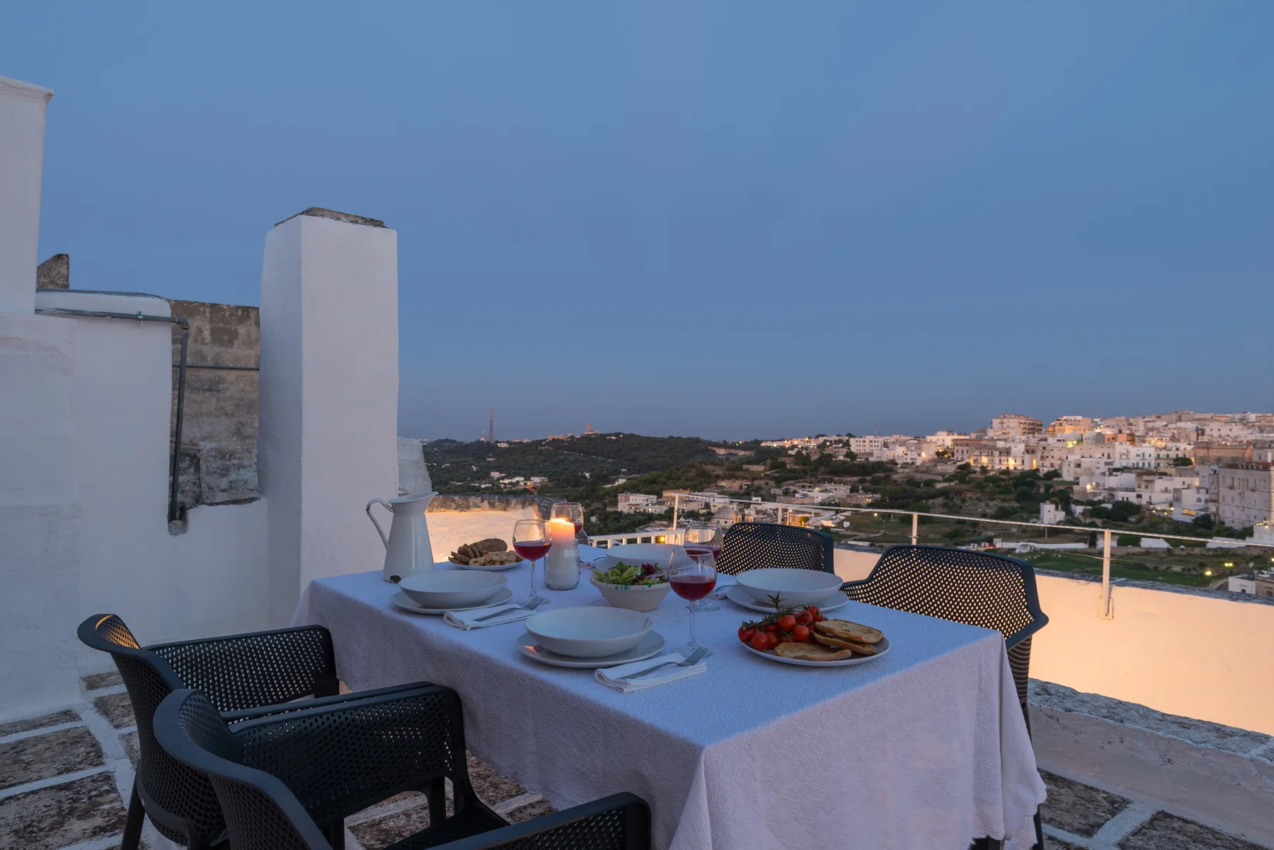 Una mesa de comedor en una terraza está puesta con platos y bebidas, frente a una localidad costera.
