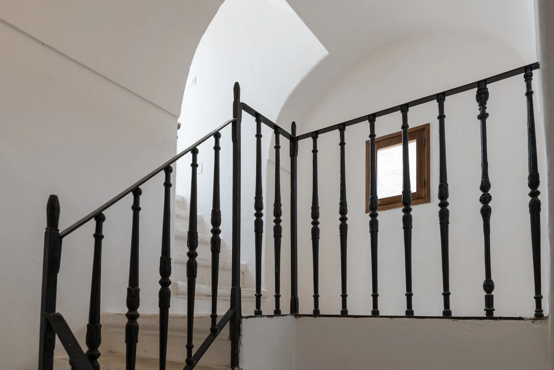 Una escalera interior blanca con una barandilla negra y una ventana pequeña.