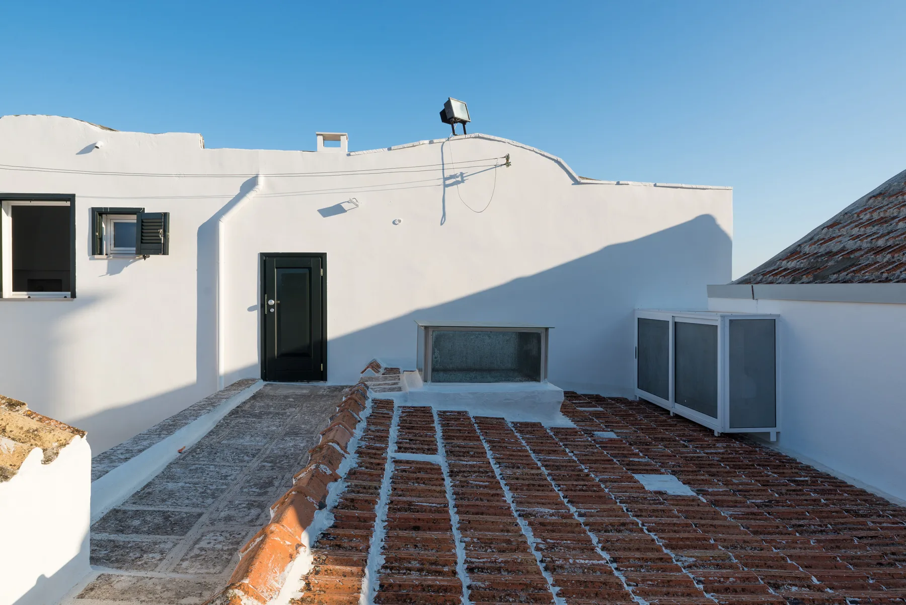 Una terraza plana en la azotea, con una pared blanca y una puerta oscura bajo un cielo despejado.