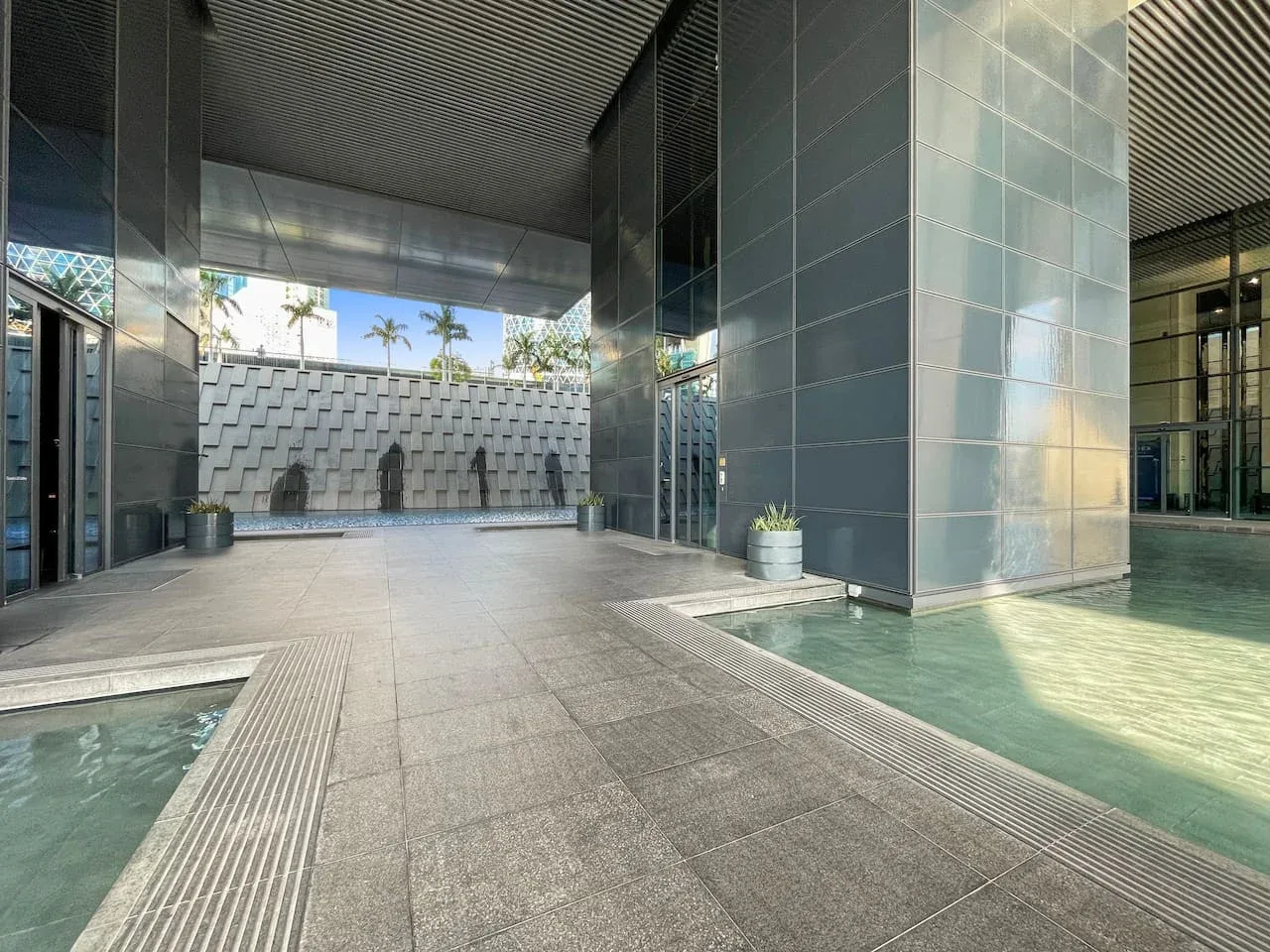 La entrada de un edificio moderno da a un amplio sendero pavimentado junto a piscinas poco profundas.