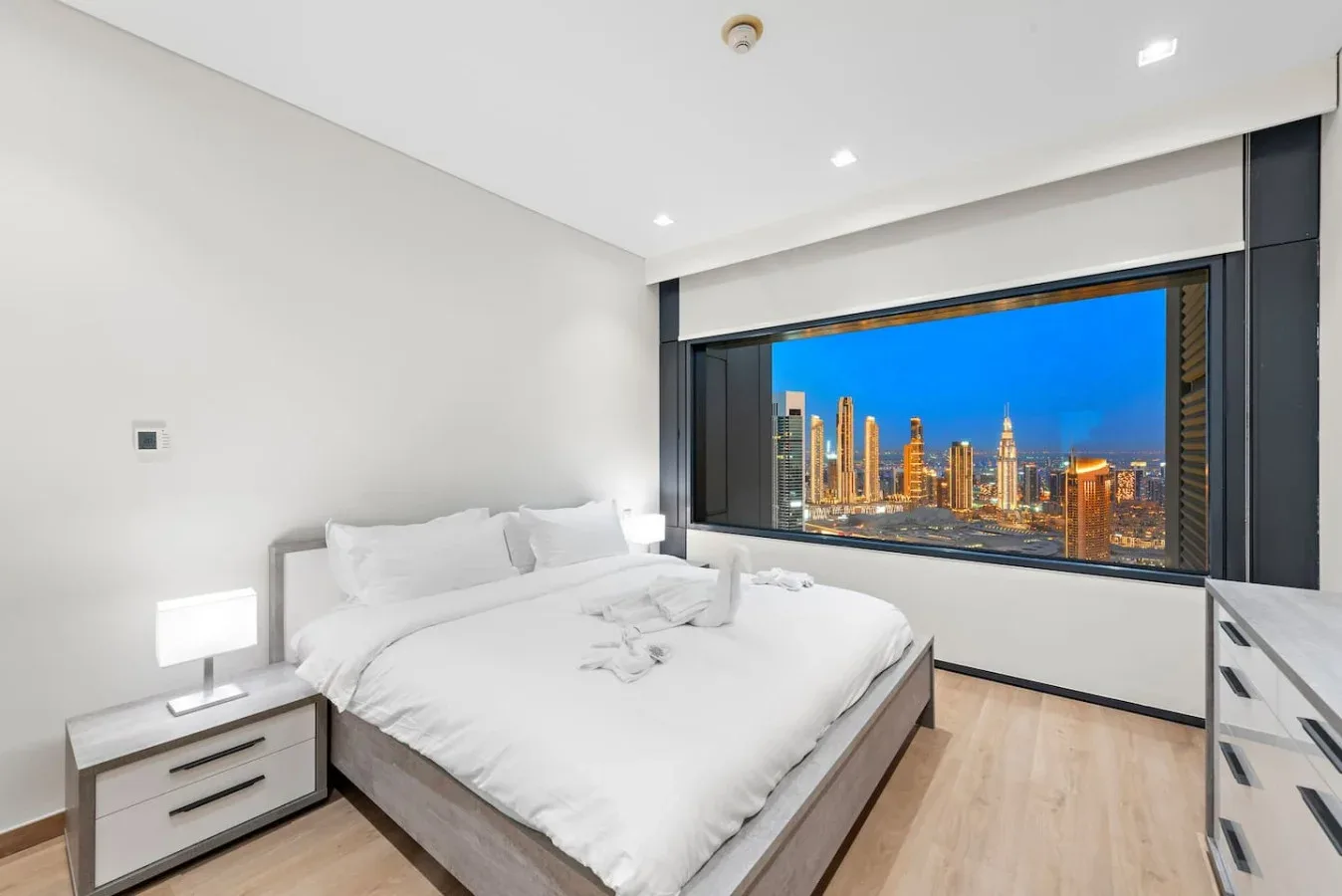 Un dormitorio luminoso con ropa de cama blanca y una gran ventana con vistas al horizonte de la ciudad.