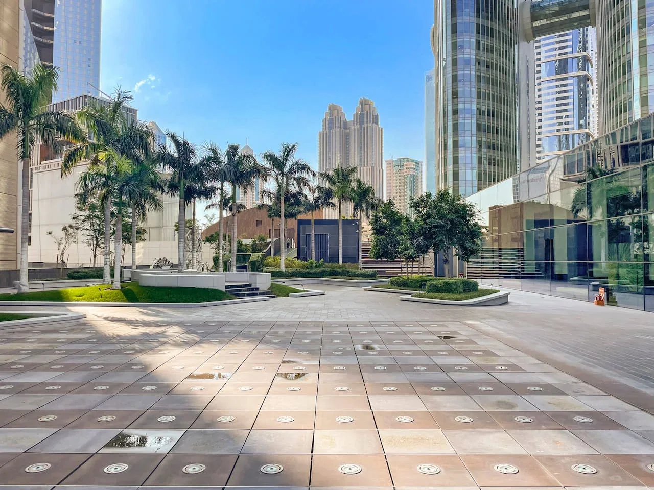 Una plaza exterior pavimentada con palmeras, jardineras y edificios altos alrededor.