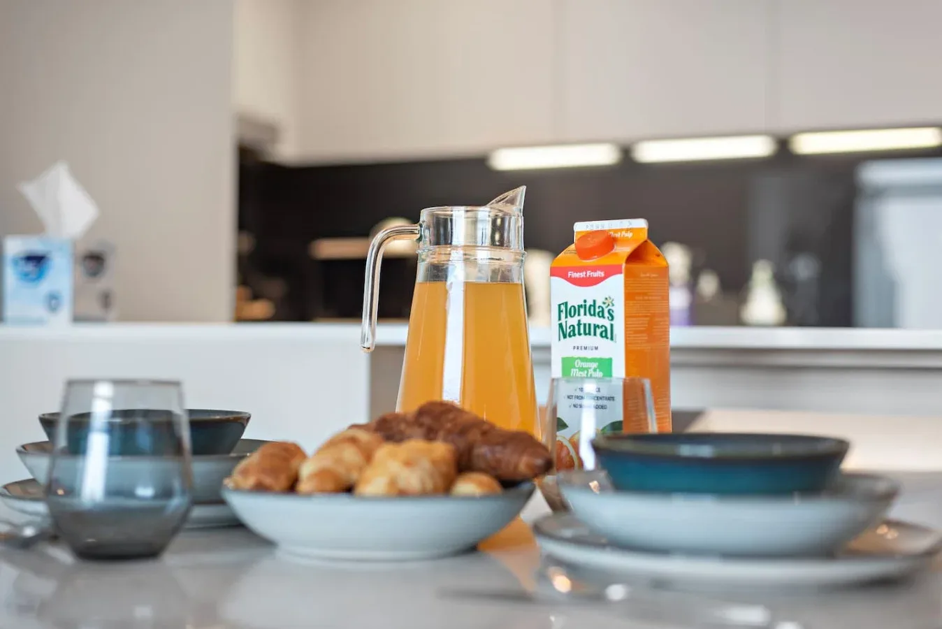 Una mesa de desayuno con una jarra de jugo, un envase de jugo de naranja, bollería y cuencos.