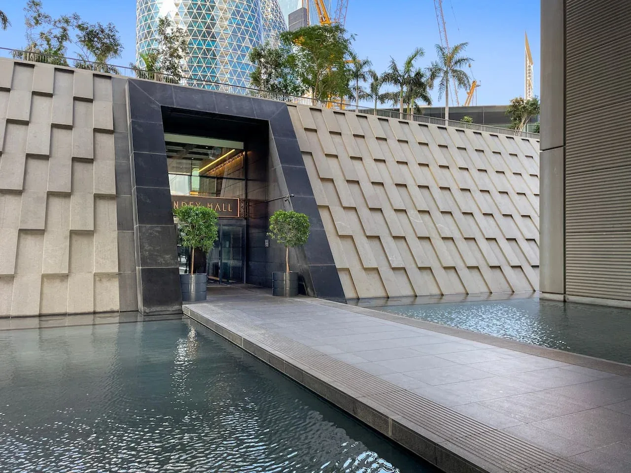 Entrada de un edificio moderno junto a una piscina reflectante, con palmeras sobre el muro.