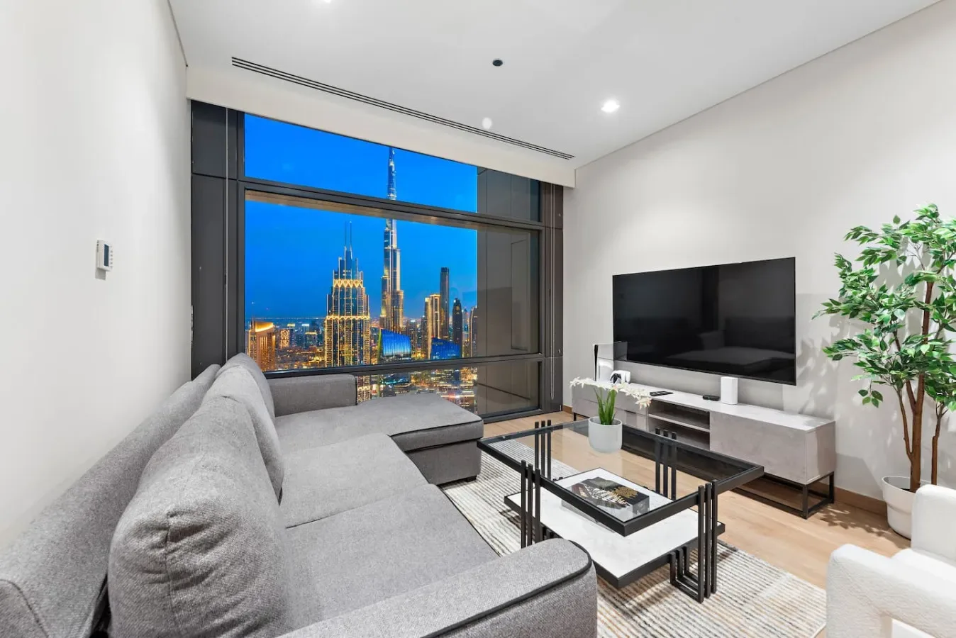 Una sala moderna con asientos grises, televisor y vistas de la ciudad a través de grandes ventanales.