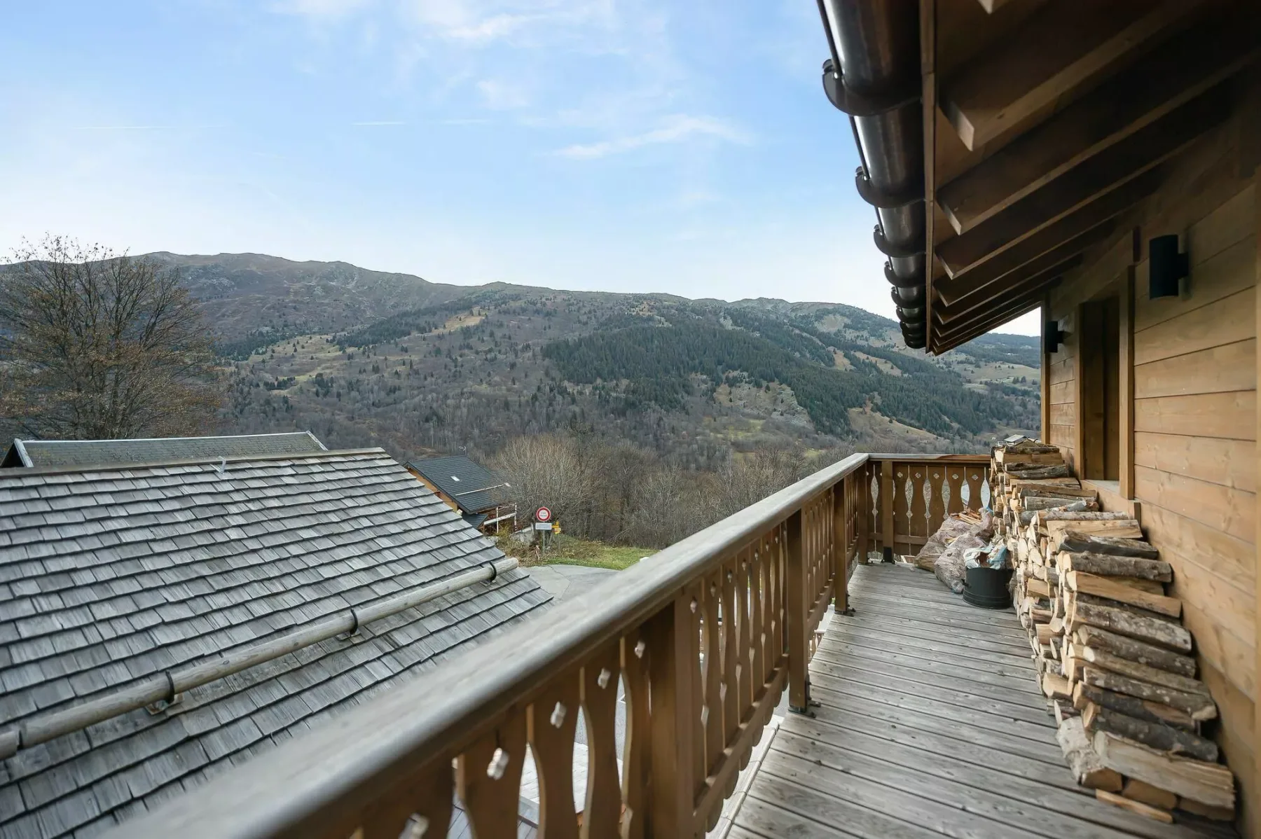 A wooden balcony overlooks a mountain landscape, with stacked firewood beside the railing.