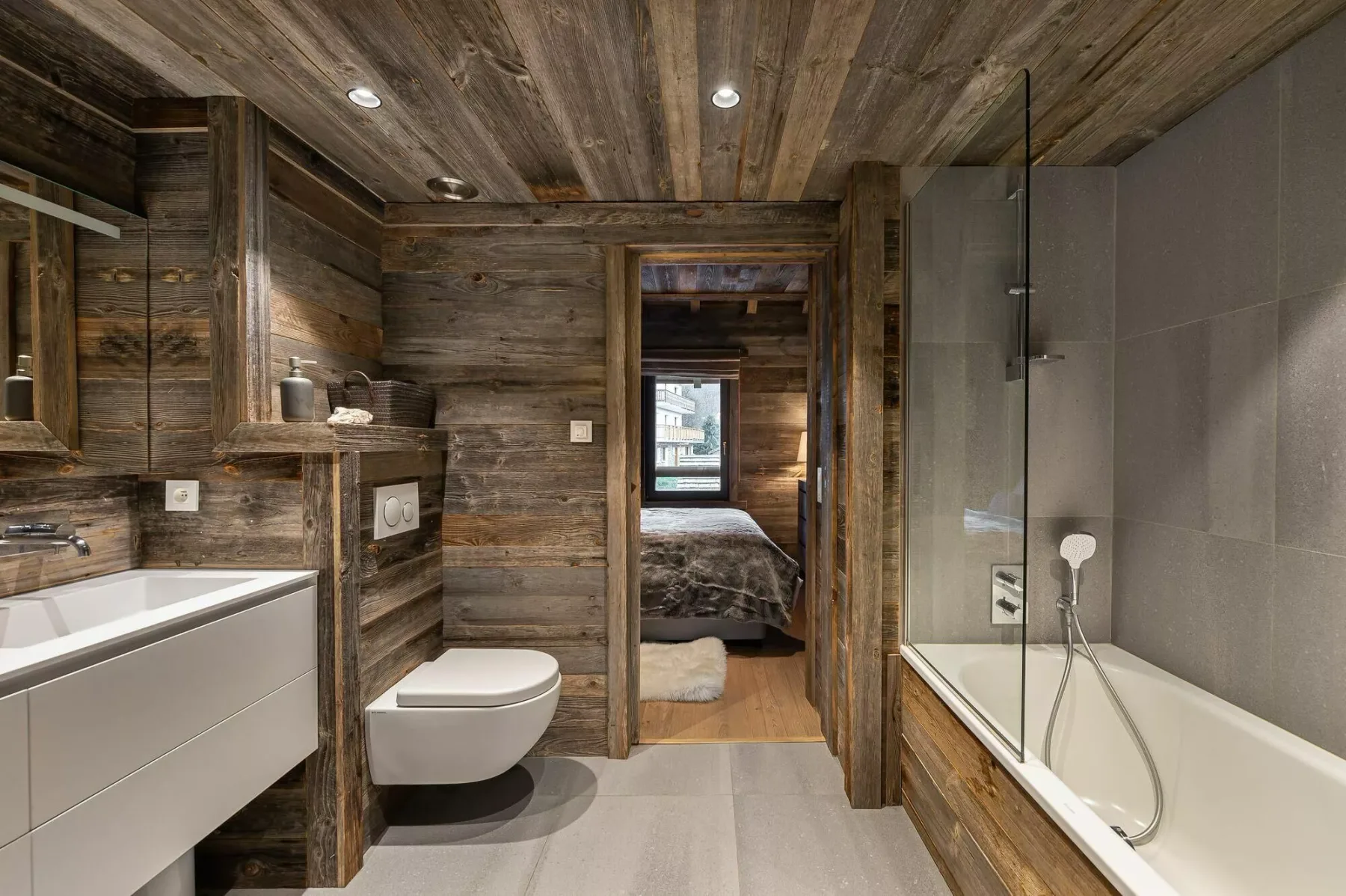 A wood-paneled bathroom with a bathtub, toilet, and vanity opens onto a bedroom.