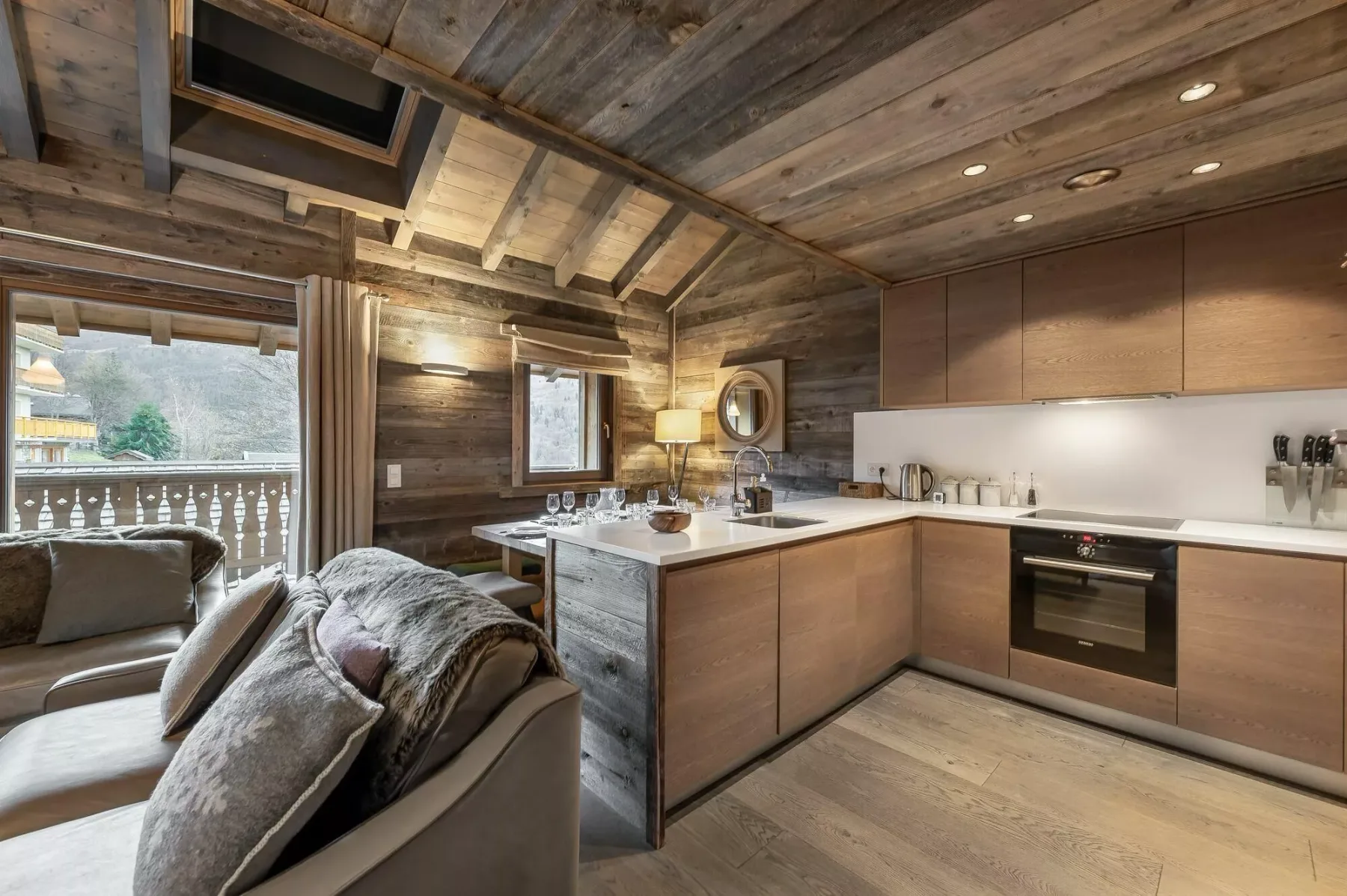 A rustic open-plan living area and kitchen with a sofa beside large windows.