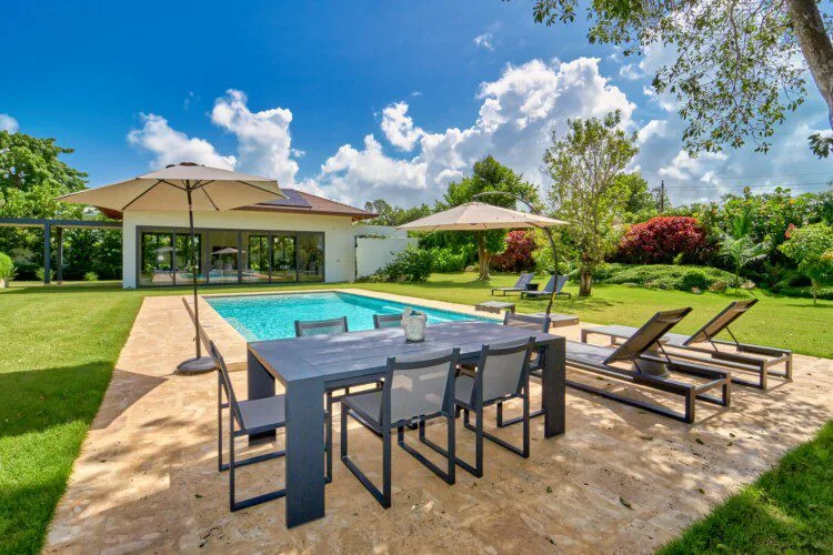 Un patio junto a la piscina tiene una mesa de comedor, tumbonas y sombrillas.