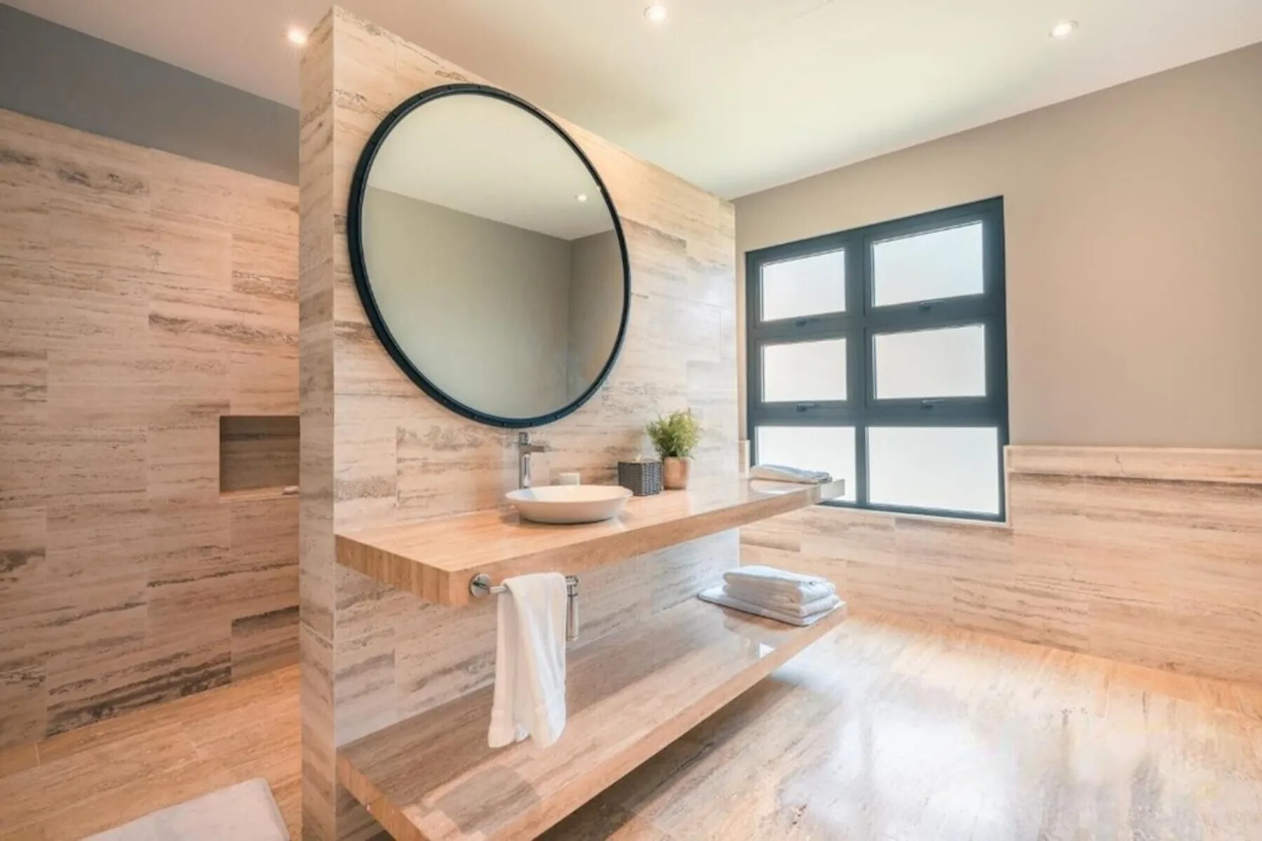 Un baño luminoso con lavabo de pared, espejo redondo y una ventana amplia.