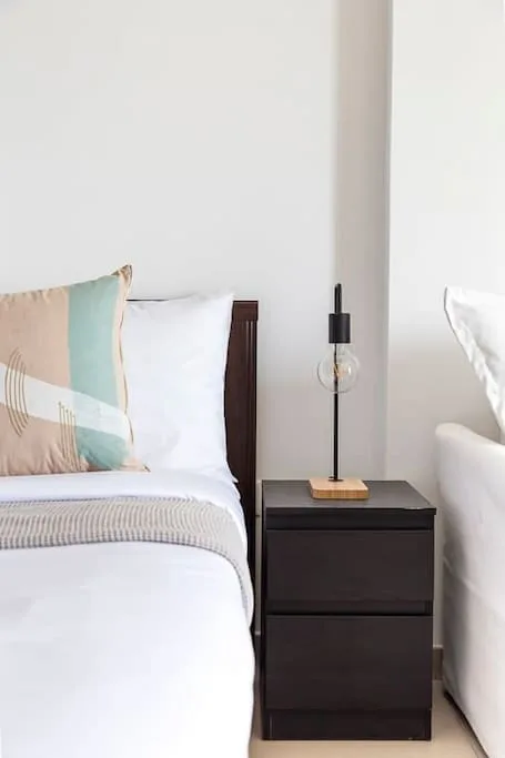 Un dormitorio luminoso con ropa de cama blanca y una mesita oscura.