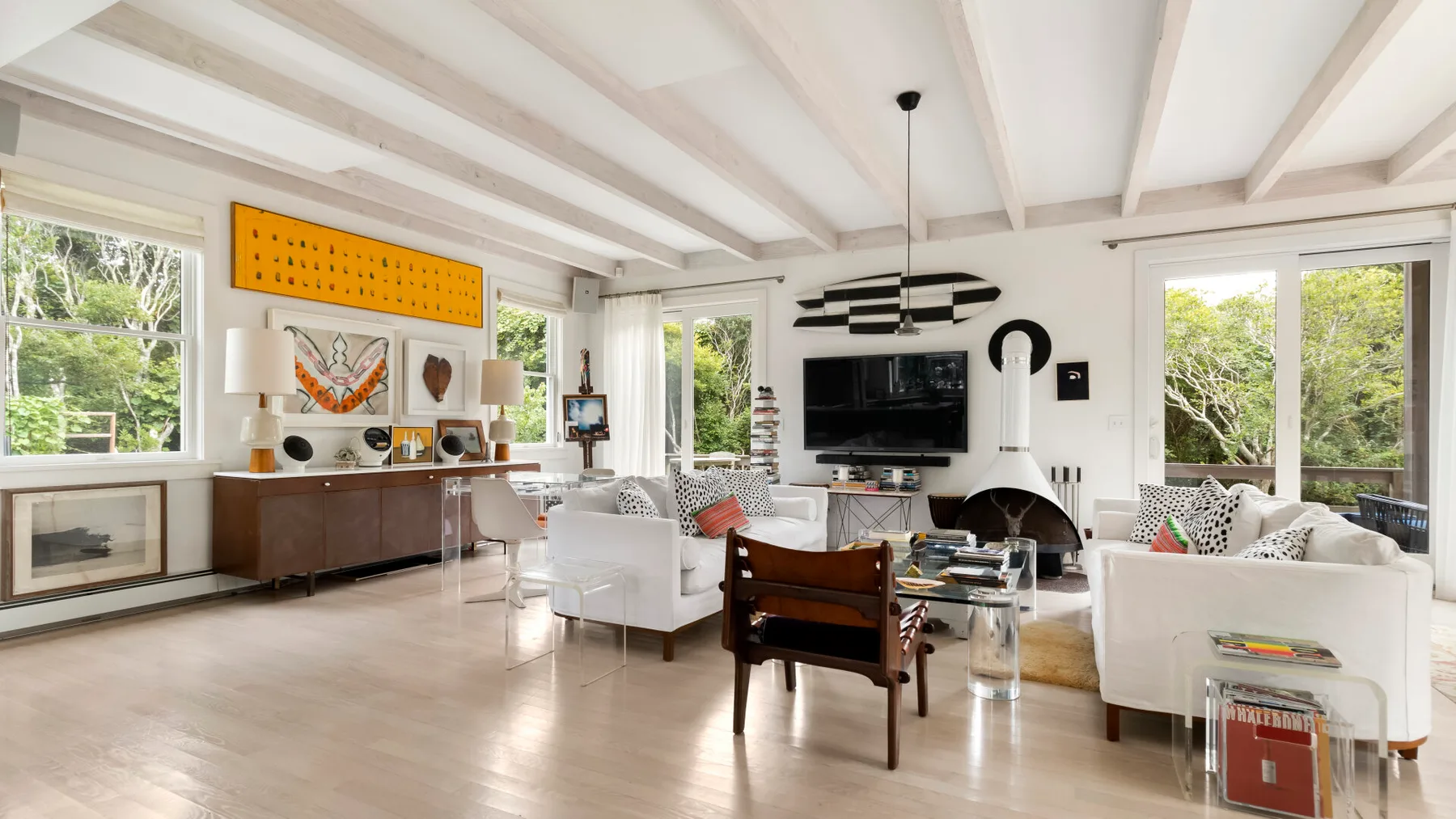 Una sala de estar luminosa con asientos blancos, un televisor y ventanas grandes.
