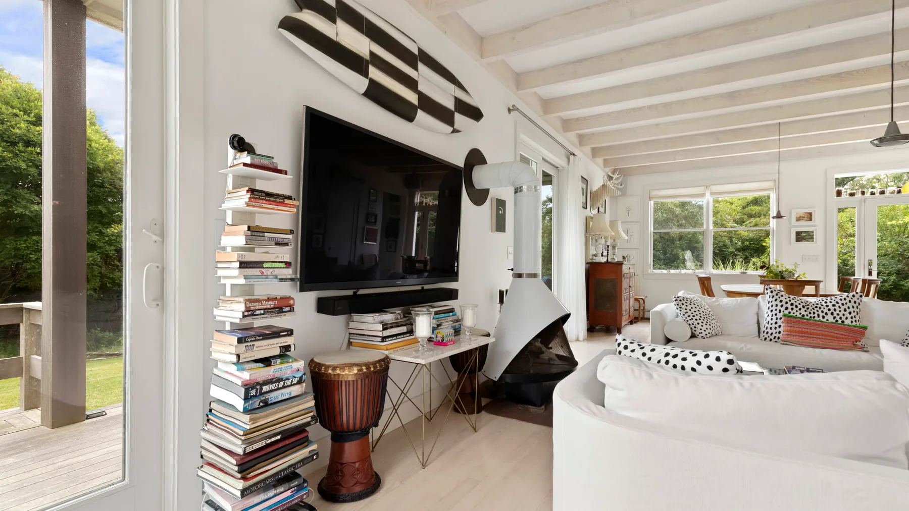 Una sala luminosa con un televisor, libros apilados y asientos blancos.