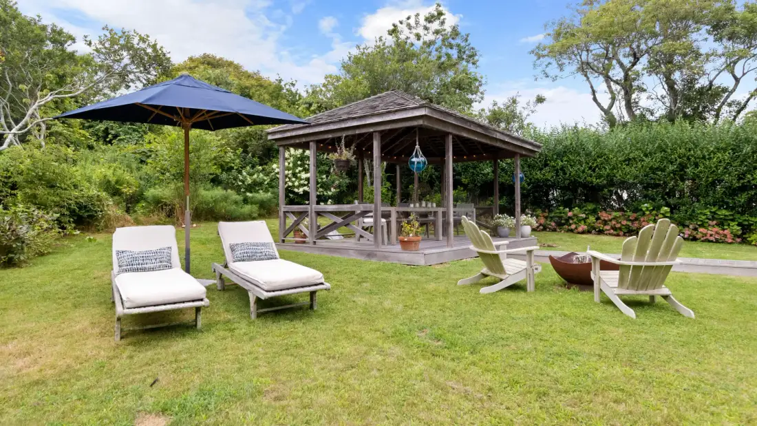 Una zona de descanso en el jardín con un pabellón de madera, sillones y una sombrilla.