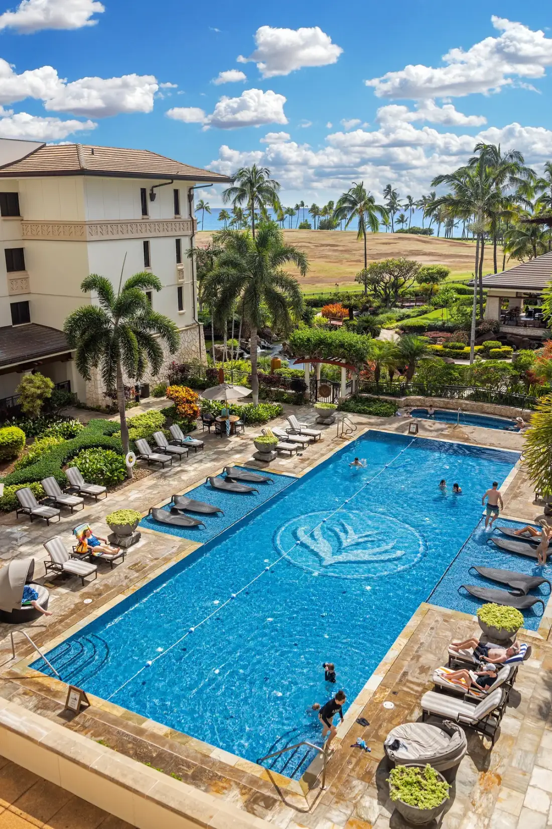 A landscaped resort pool area with lounge seating beneath a bright, cloudy sky.