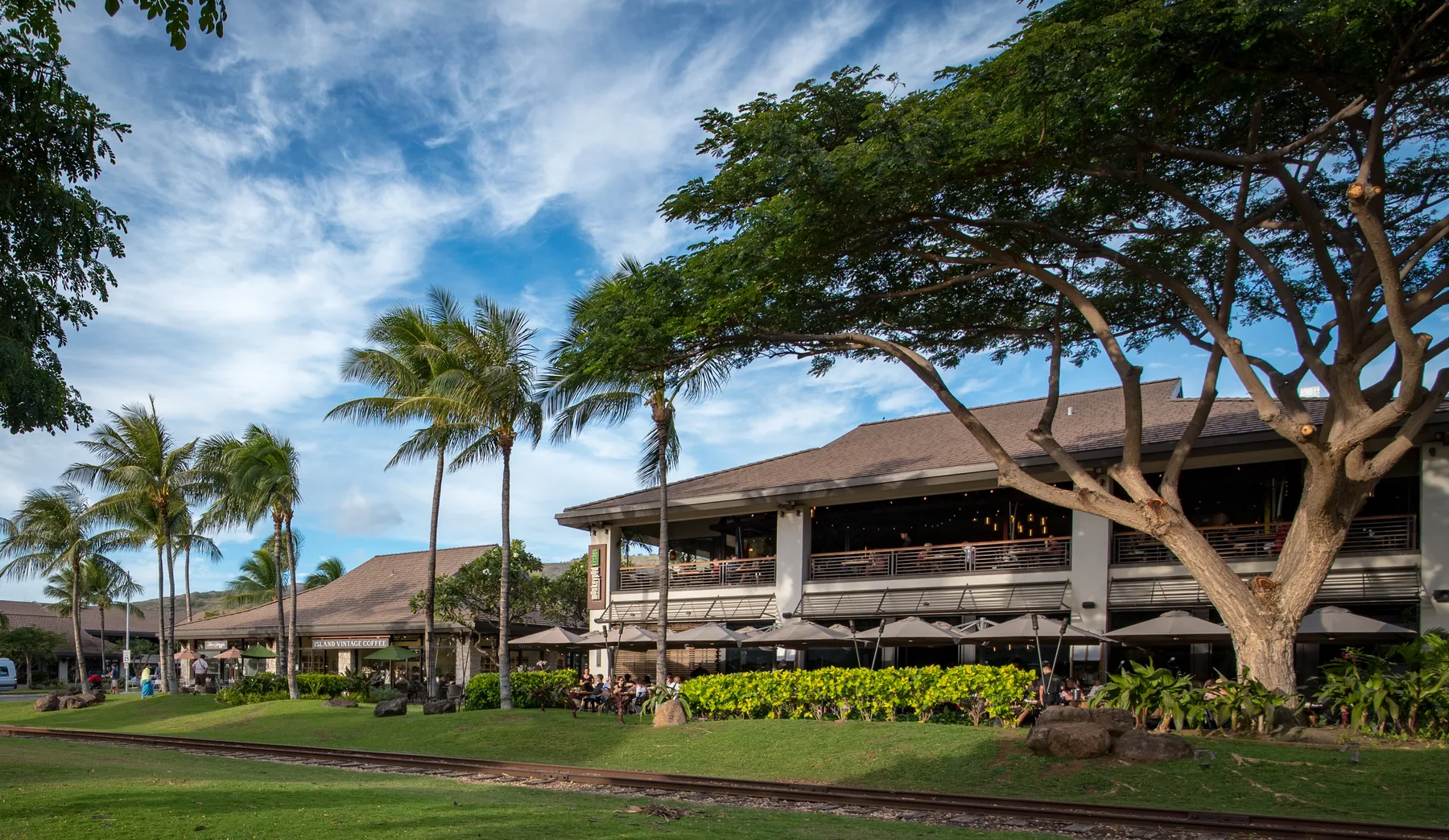 A low-rise open-sided building stands beside a landscaped lawn and palm trees.