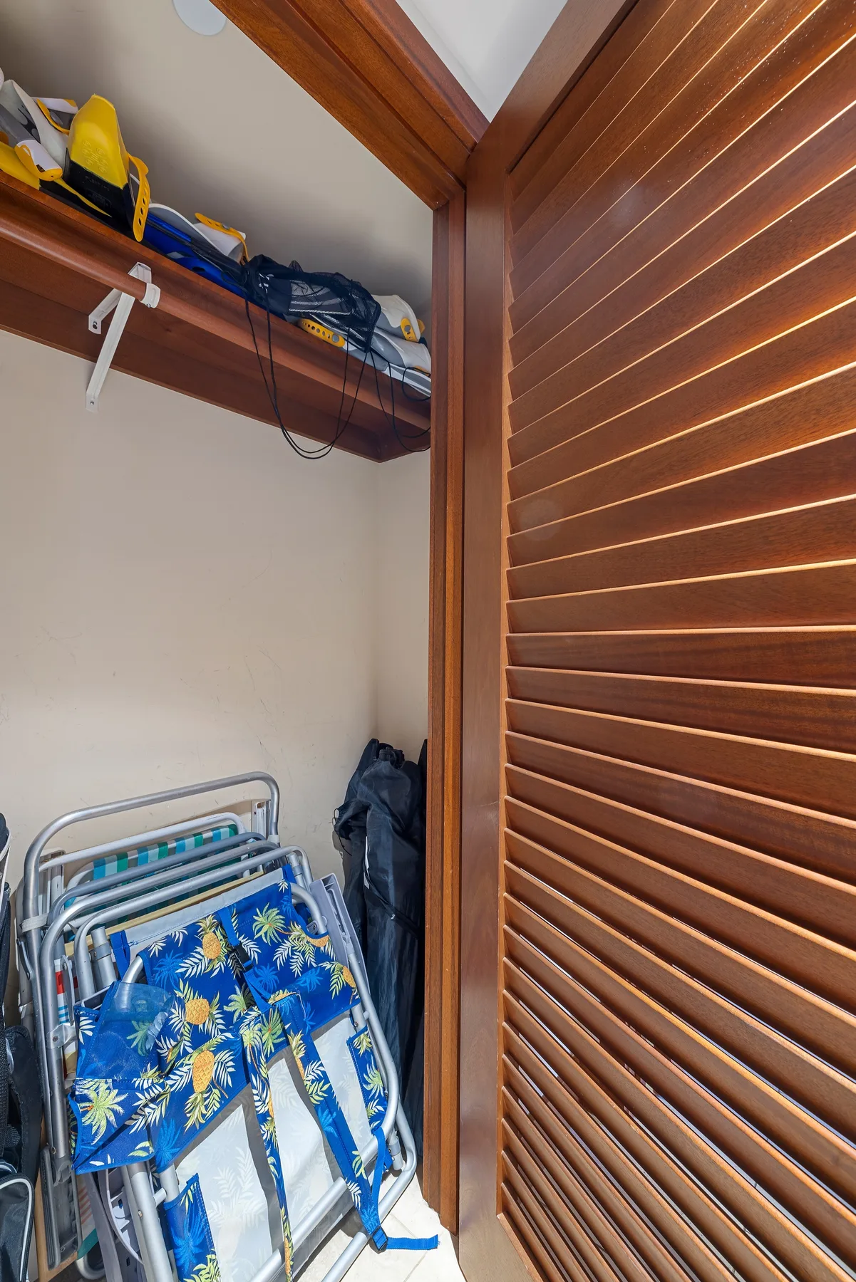 A storage area with folded beach chairs and bags on a shelf beside wooden louvered doors.