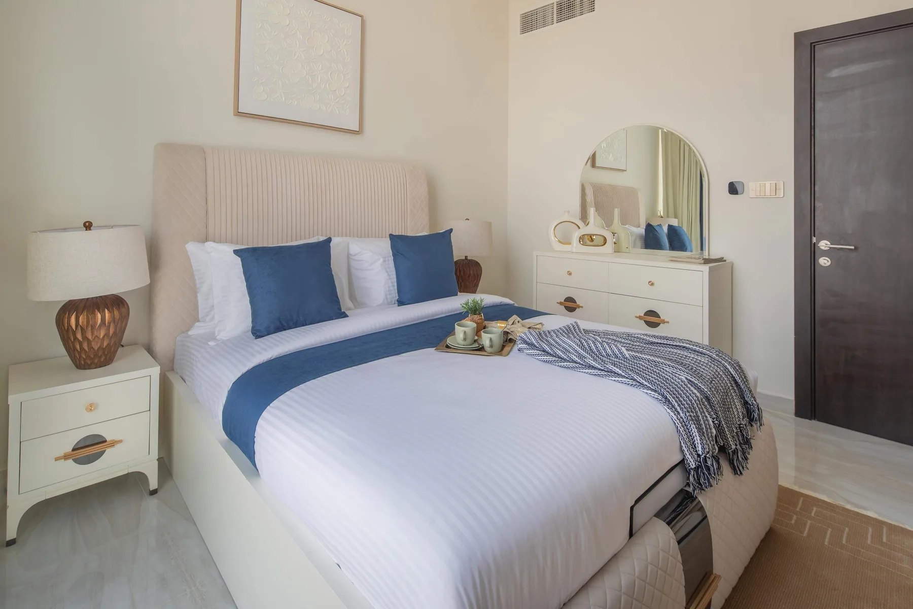 Un dormitorio con una cama tendida, almohadas azules y una cómoda con espejo.