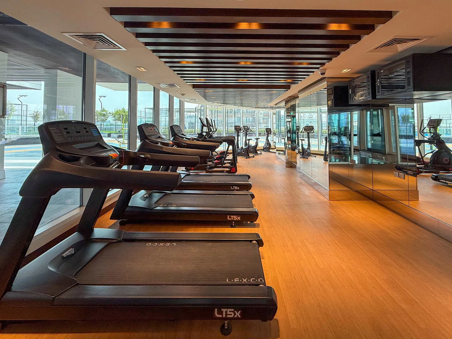Un gimnasio luminoso con cintas de correr y otros equipos de ejercicio junto a grandes ventanales.