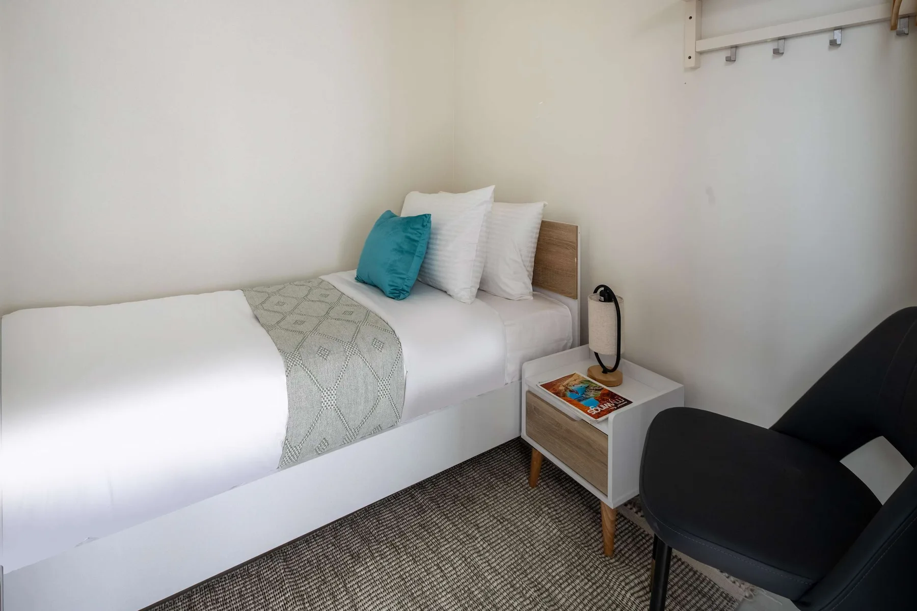 Una cama individual con ropa de cama blanca y una mesita está junto a una silla oscura.