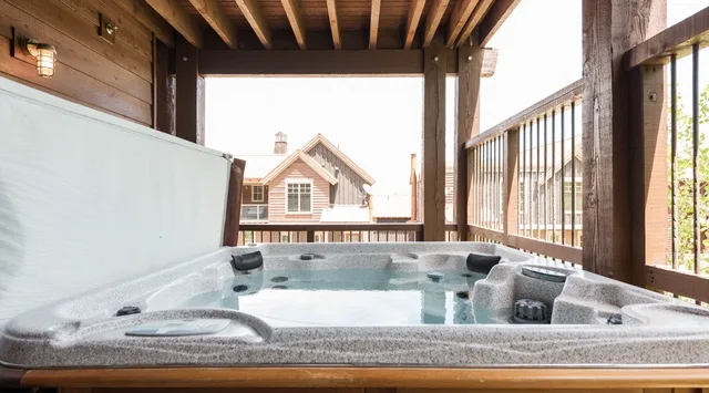 Un jacuzzi está bajo una terraza de madera cubierta, con edificios vecinos al fondo.