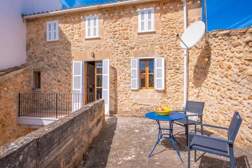 A stone house opens onto a sunny terrace with a table and seating.
