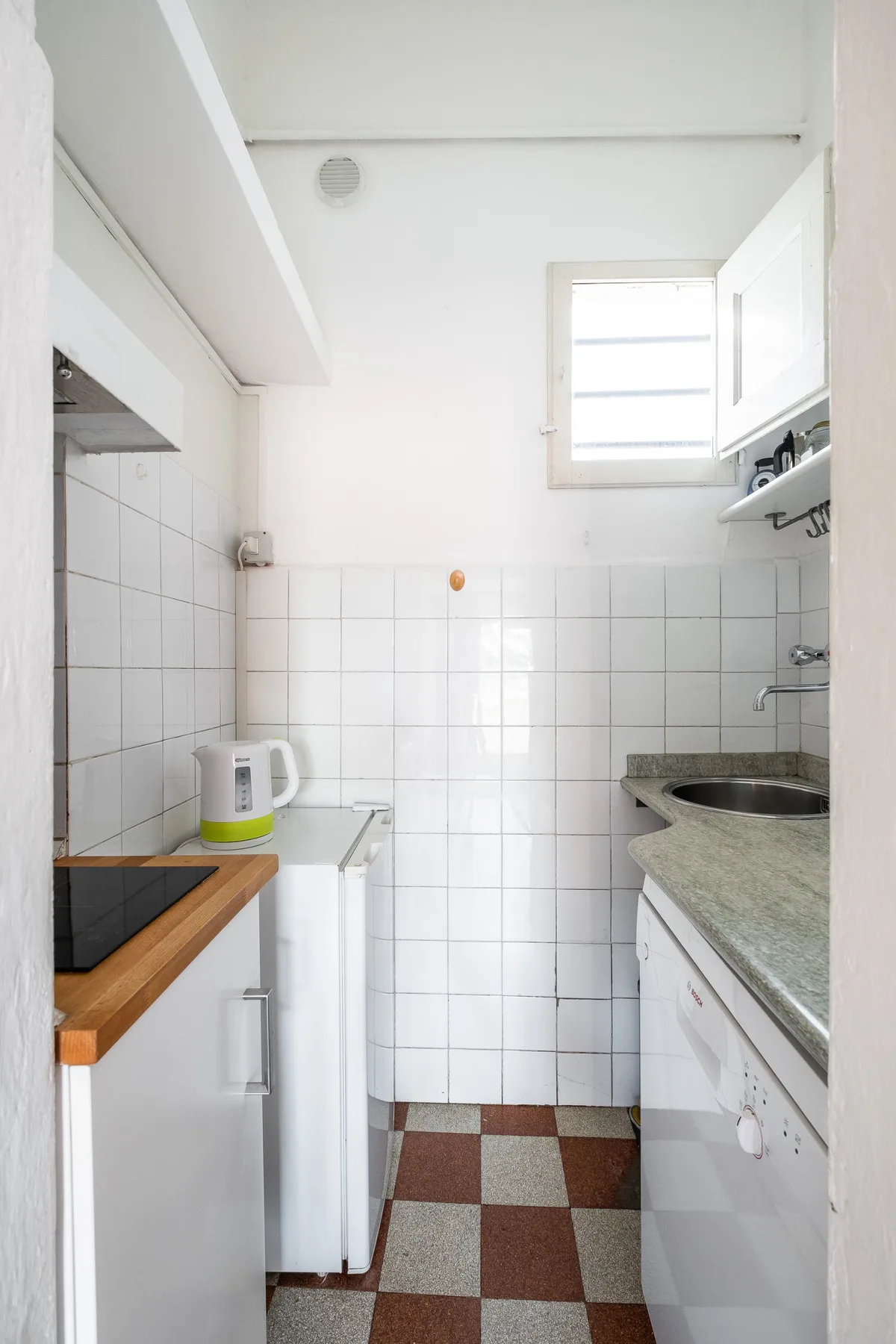 A narrow kitchen with white tiled walls, counters, and a small window.