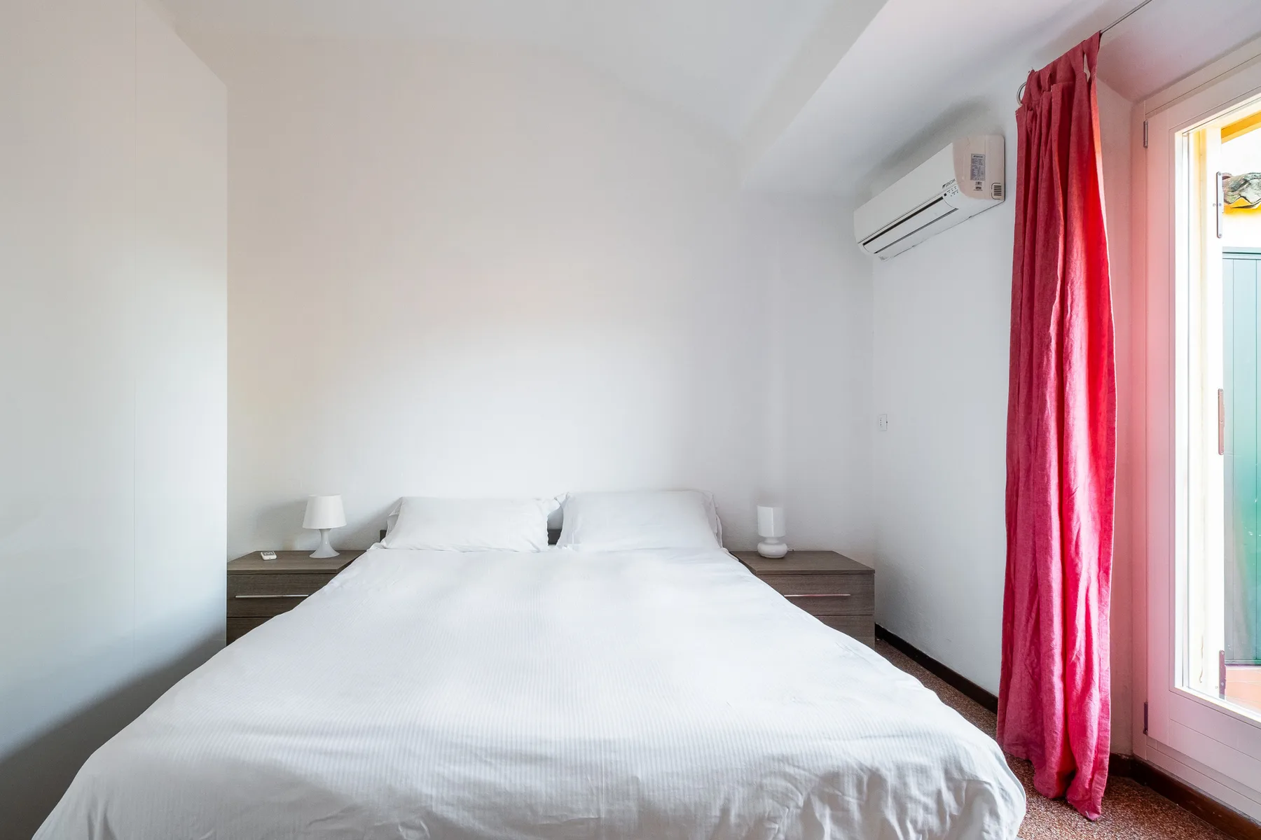 A bright bedroom with a neatly made bed and red curtains beside the window.