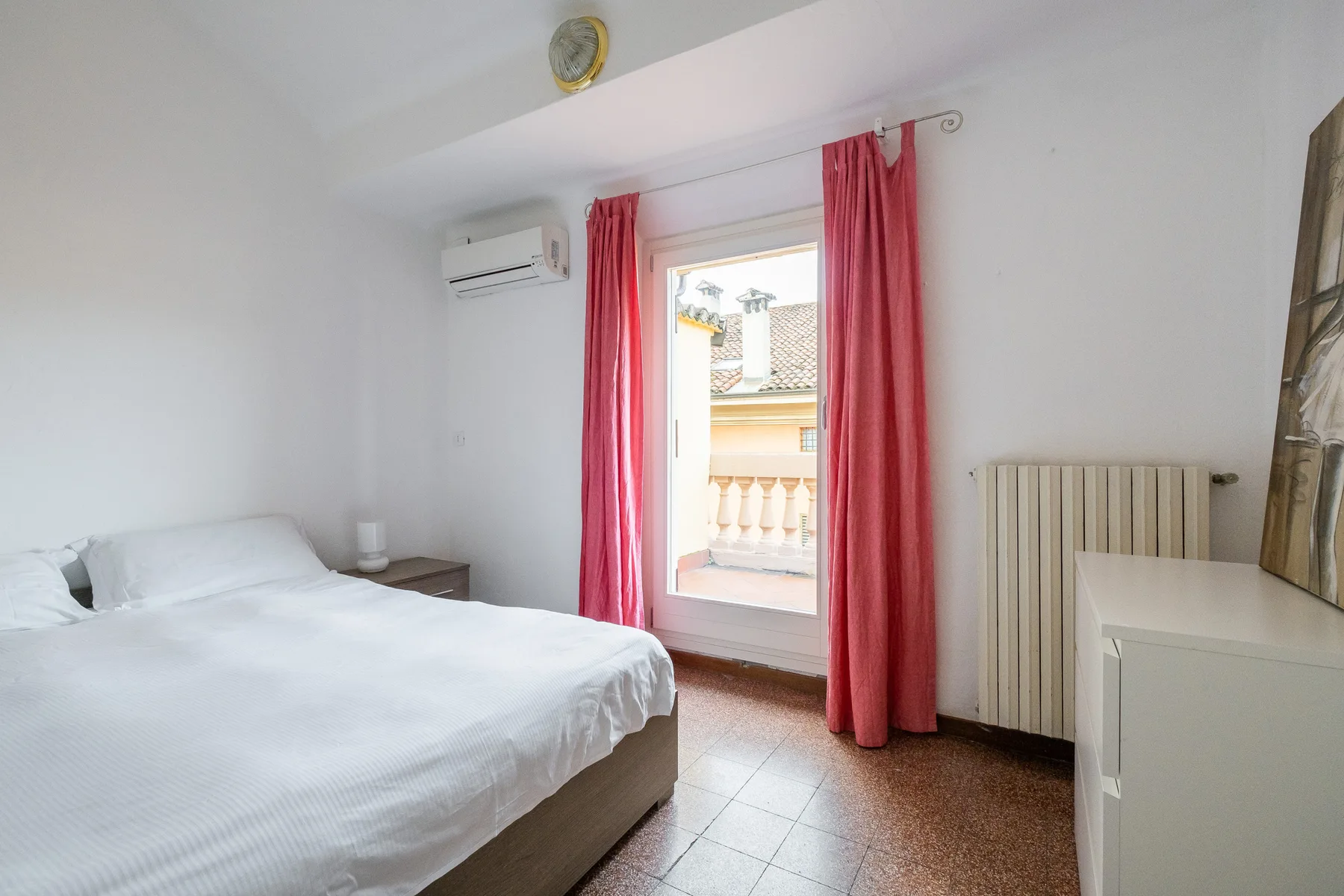 Un dormitorio luminoso con una cama, cortinas rojas y una puerta al balcón.