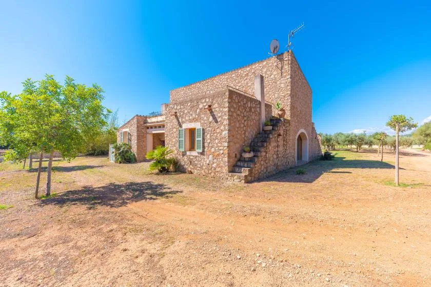 A stone building stands in a sunny, dry yard with trees nearby.