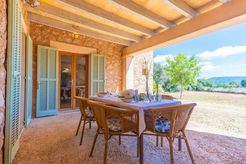 A covered outdoor dining area has a table and chairs beside shuttered glass doors.