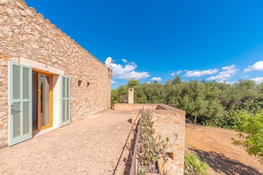 A stone house opens onto a sunlit terrace overlooking green trees.