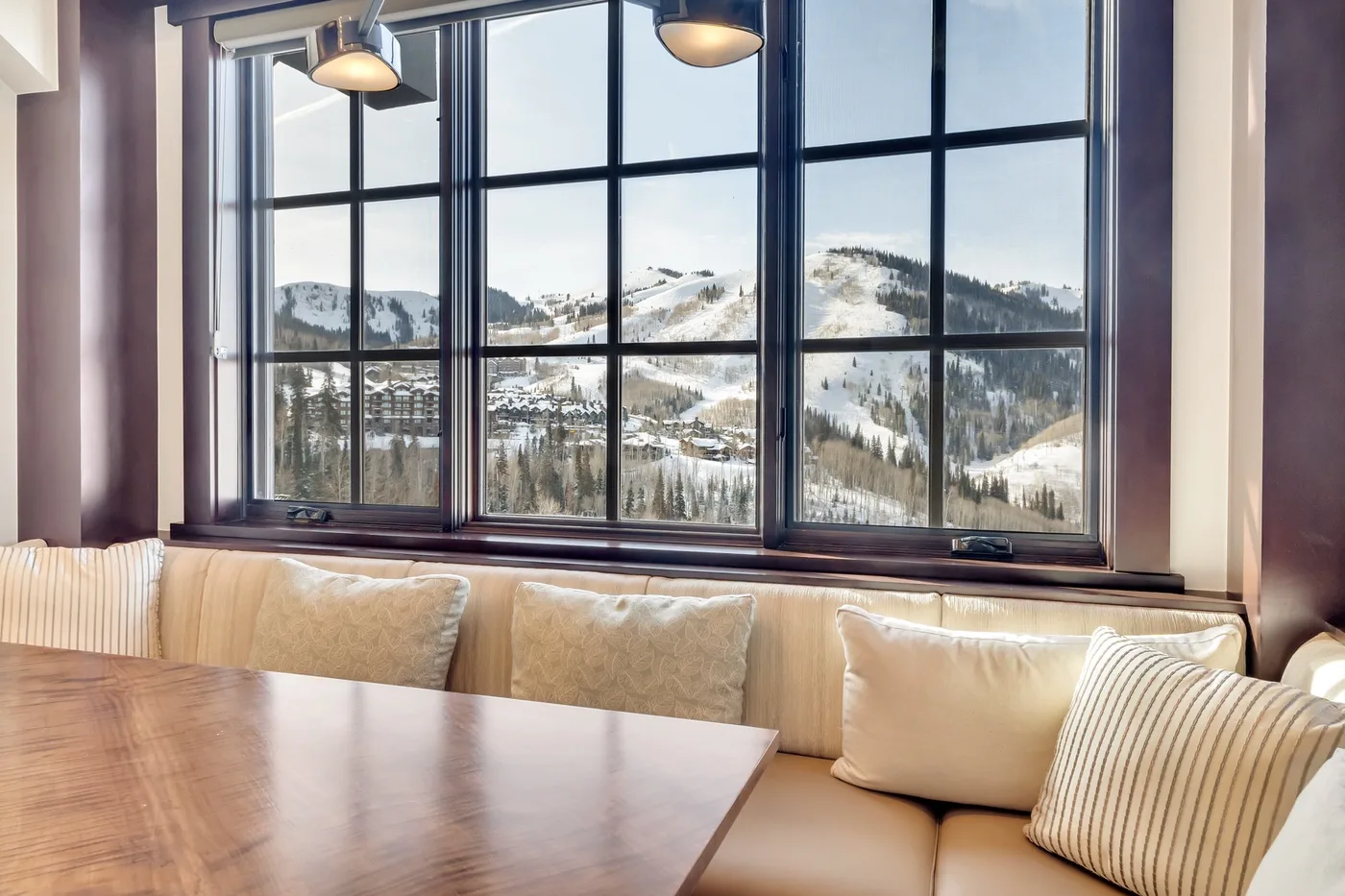 A dining table and cushioned seating sit beside a large window overlooking snowy mountains.
