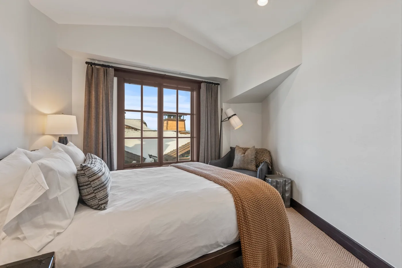 A bedroom with a neatly made bed and a large window overlooking nearby rooftops.