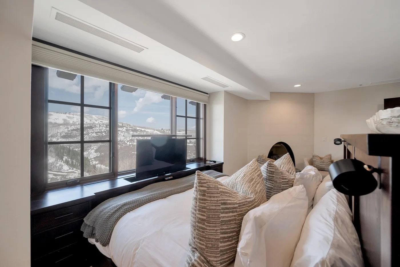 A bedroom with a bed, layered pillows, and a large window overlooking the city.