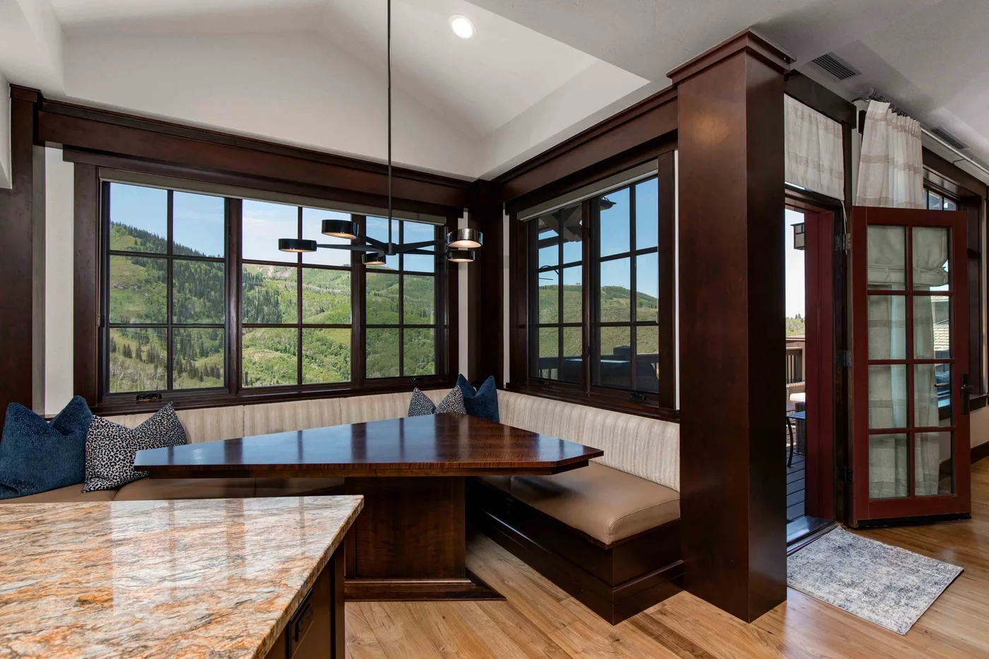 A dining nook with a built-in bench sits beside large windows overlooking green hills.