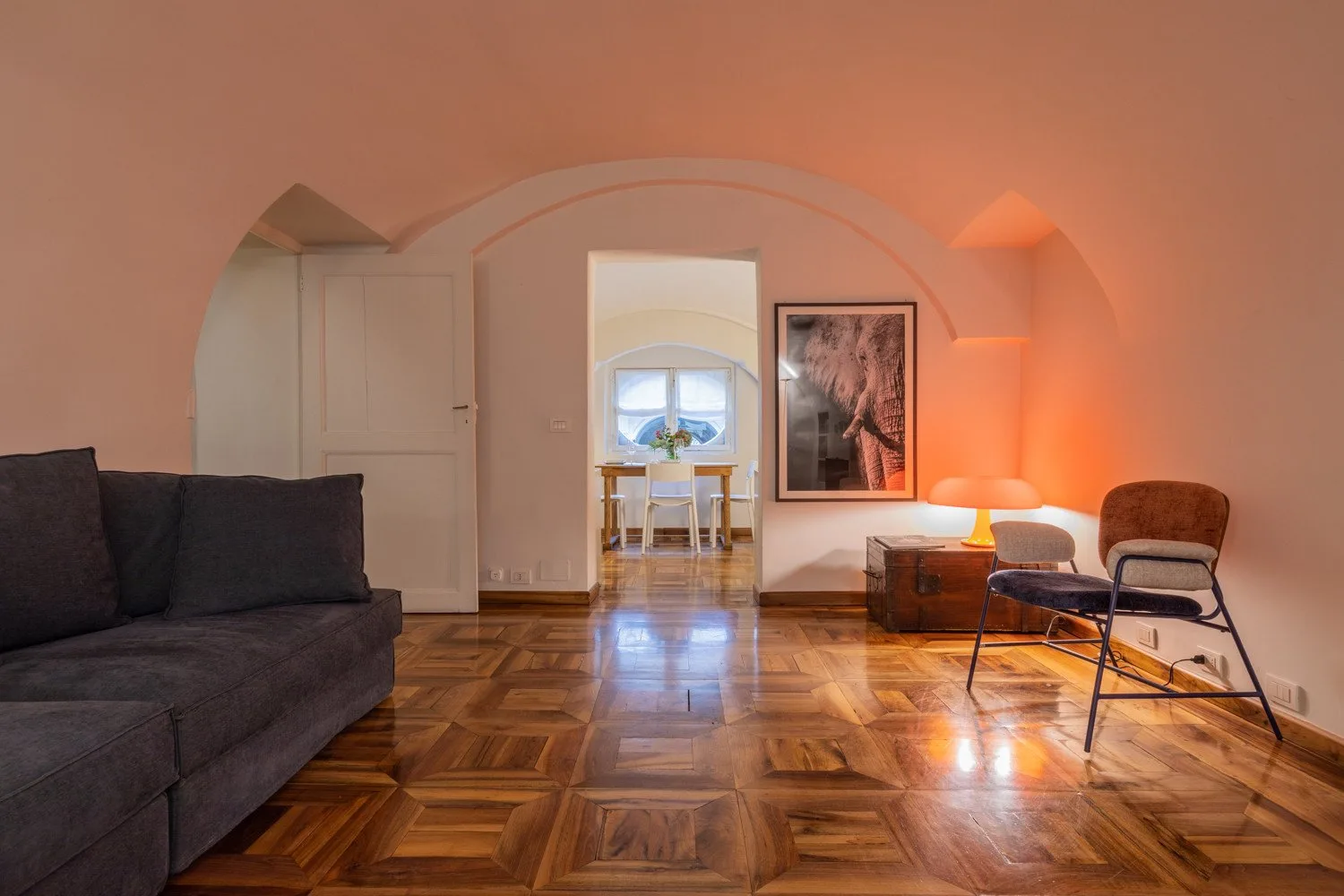 Una sala de estar con asientos oscuros, suelo de parquet brillante y un arco.
