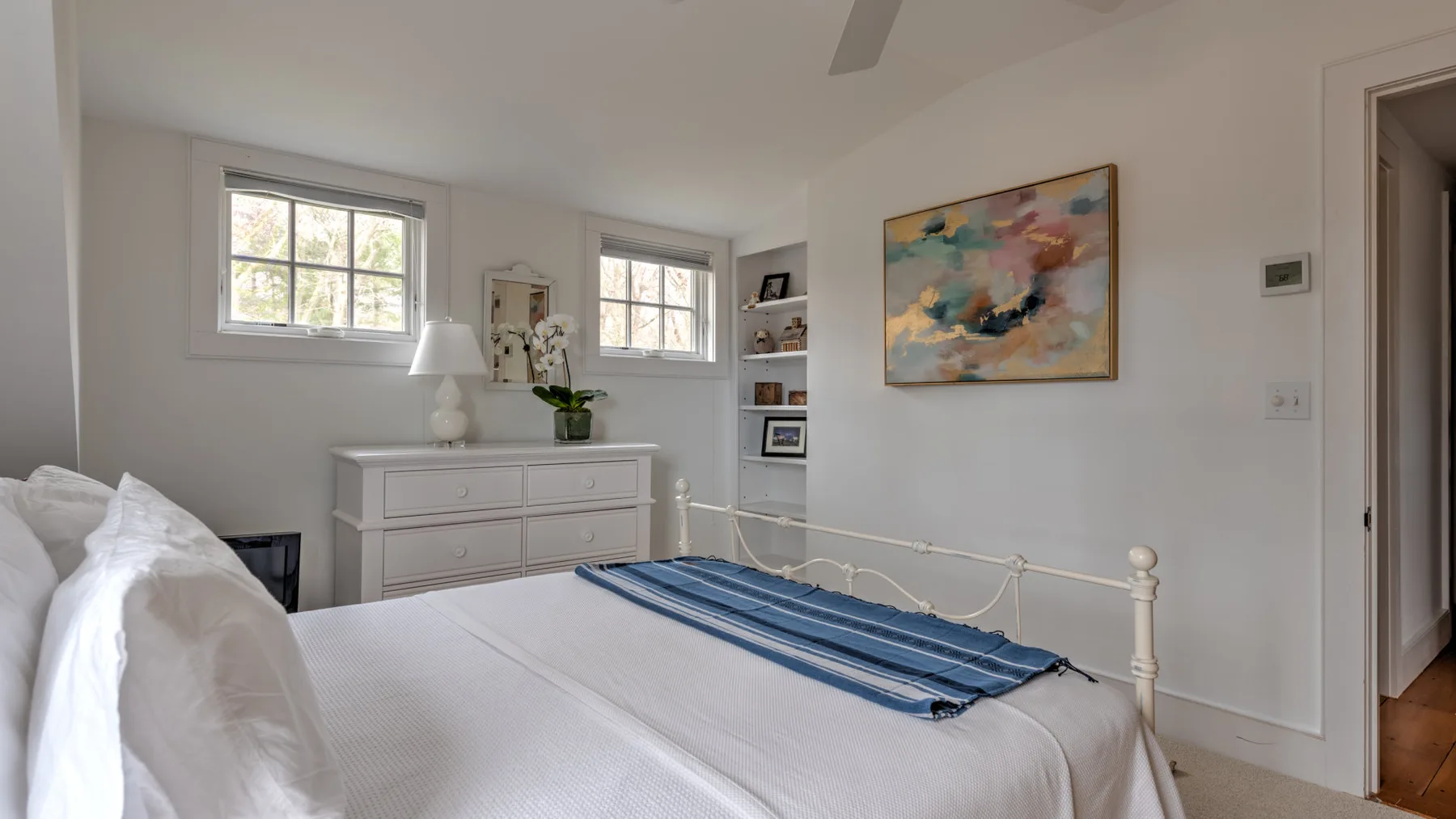 Un dormitorio luminoso con una cama blanca, una manta azul a rayas y arte abstracto en la pared.