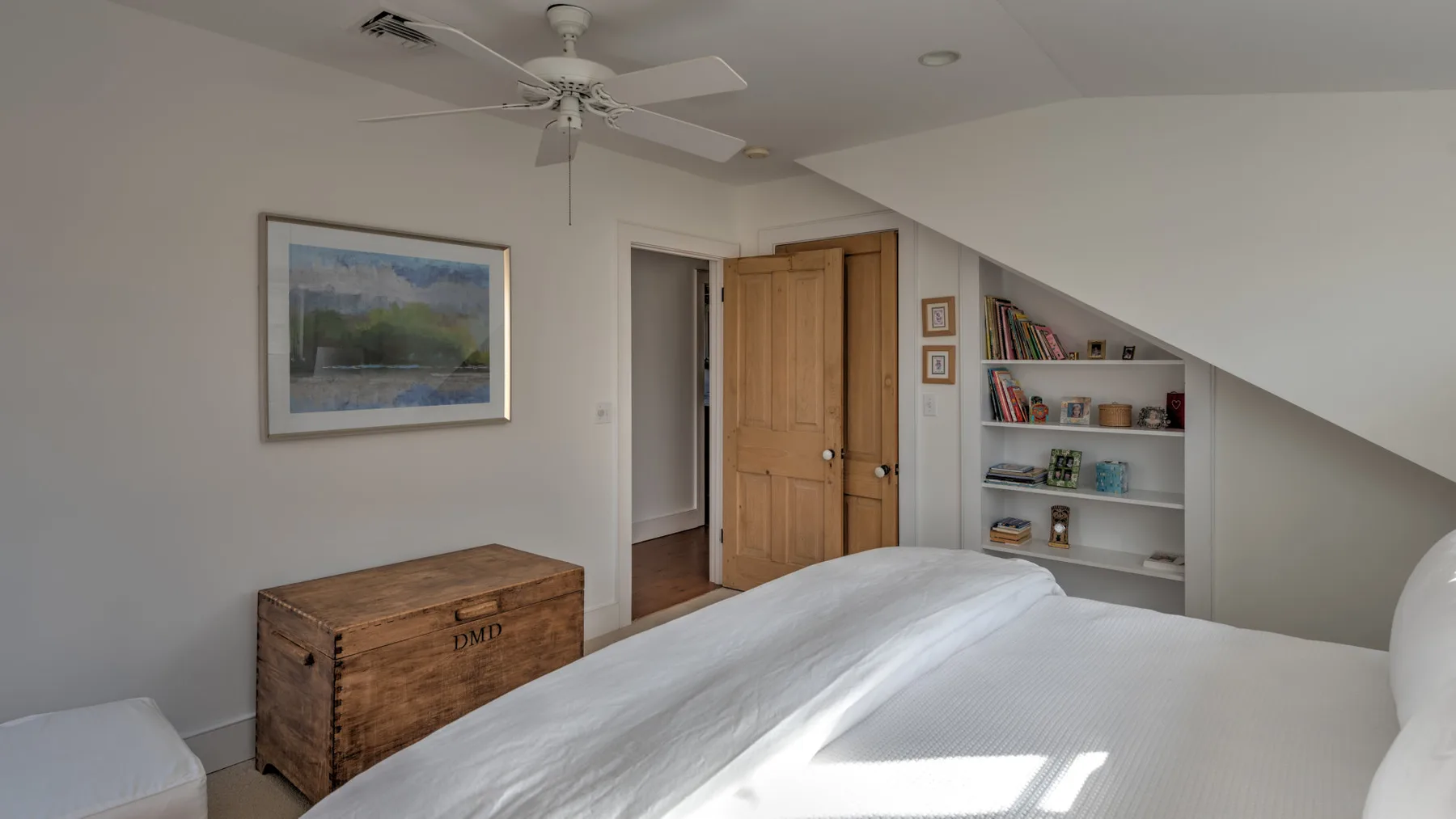 Un dormitorio con una cama, un baúl de madera y estantes bajo un techo inclinado.