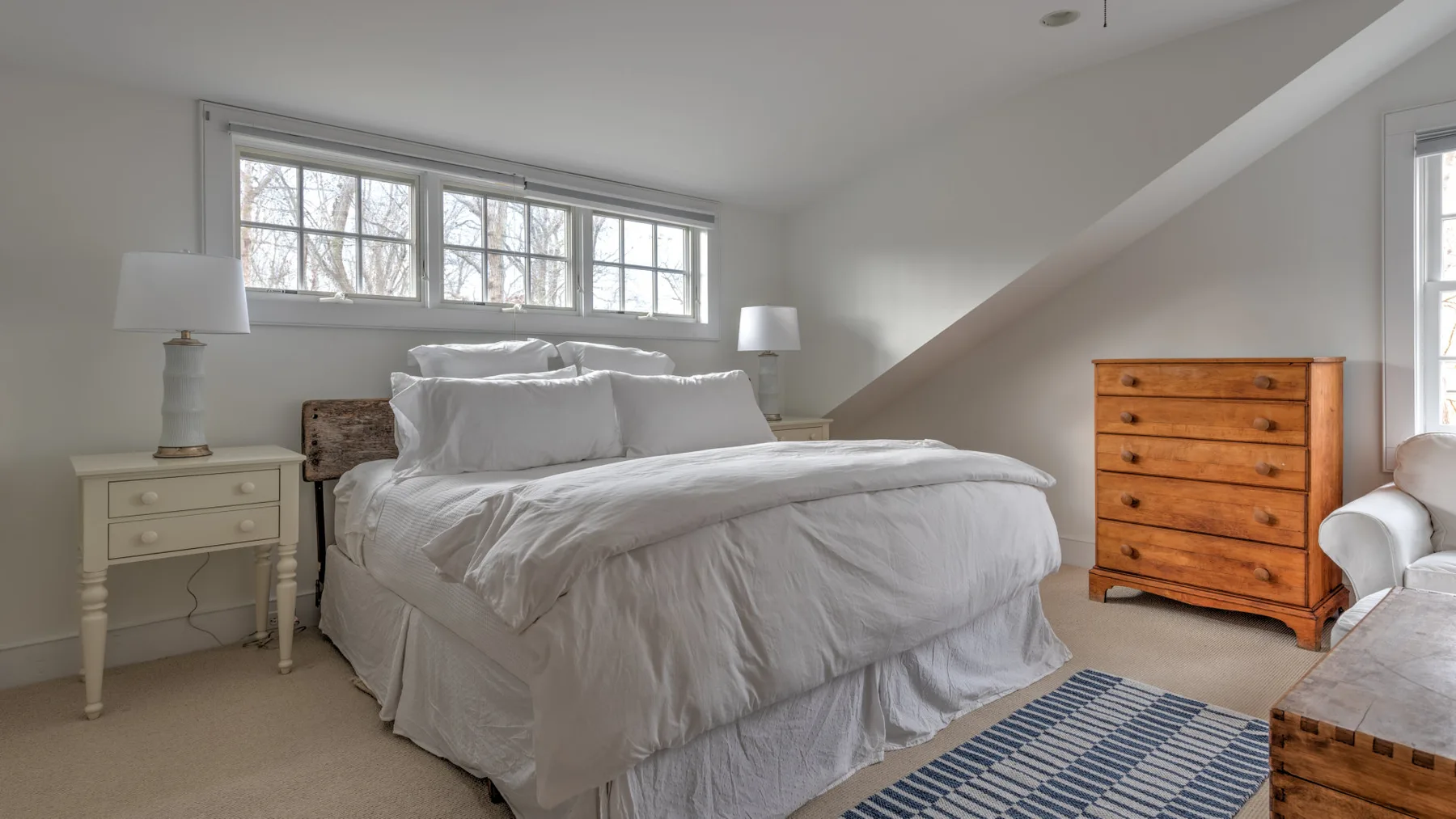 Un dormitorio luminoso con una cama bajo una fila de ventanas y muebles de madera.