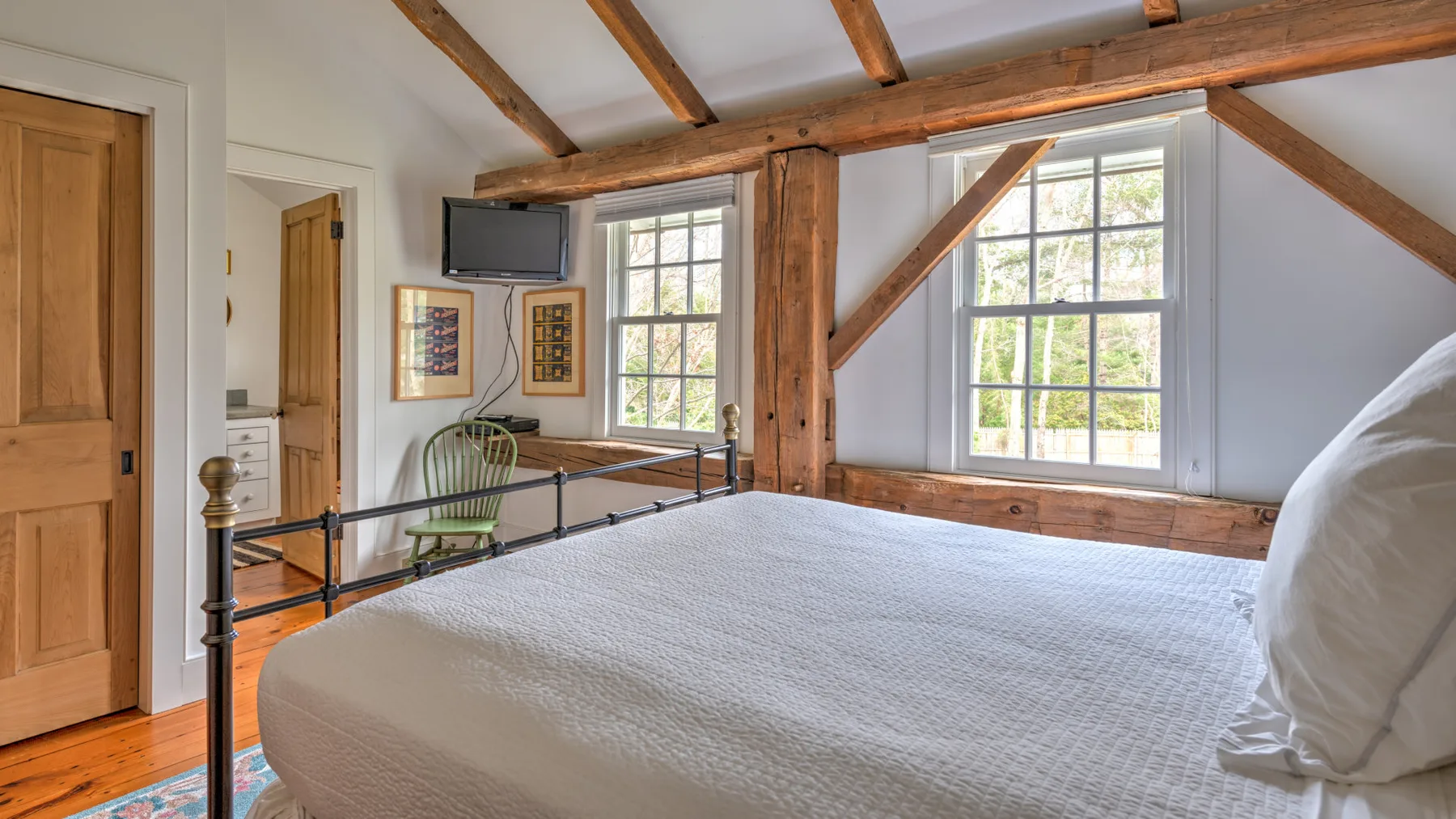 Dormitorio con una cama grande bajo vigas de madera vistas y ventanas luminosas.