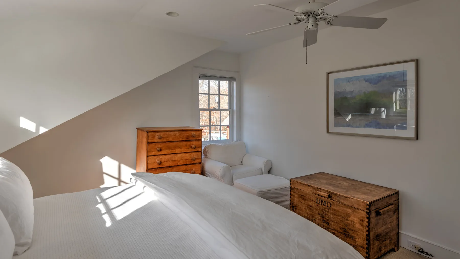 Un dormitorio luminoso con cama, arcón de madera y asientos bajo un techo inclinado.